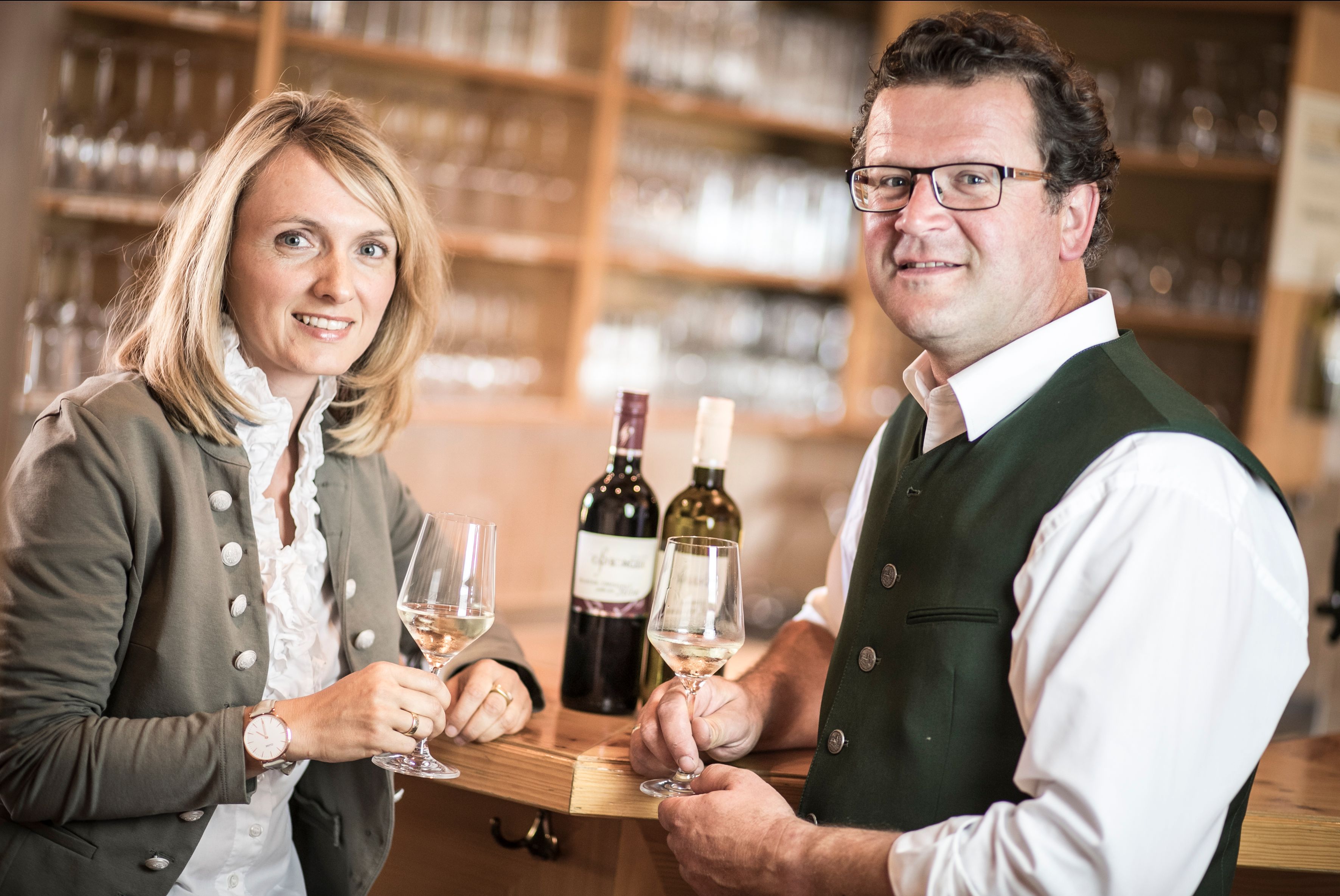 Zwei Personen in traditioneller Kleidung halten Weingläser in einem Heurigenlokal.