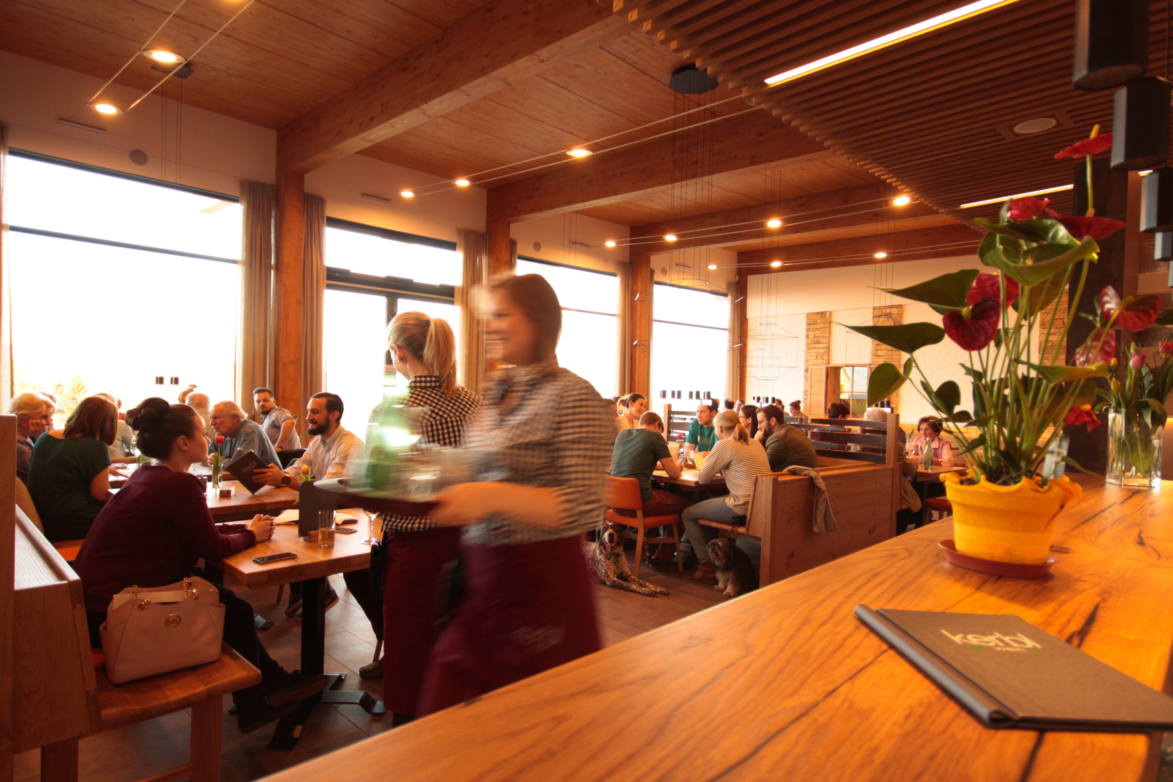 Ein gut besuchter Gastraum mit Holzmöbeln, Gästen an Tischen und einer Bedienung im Vordergrund. Eine Pflanze steht auf der Theke.