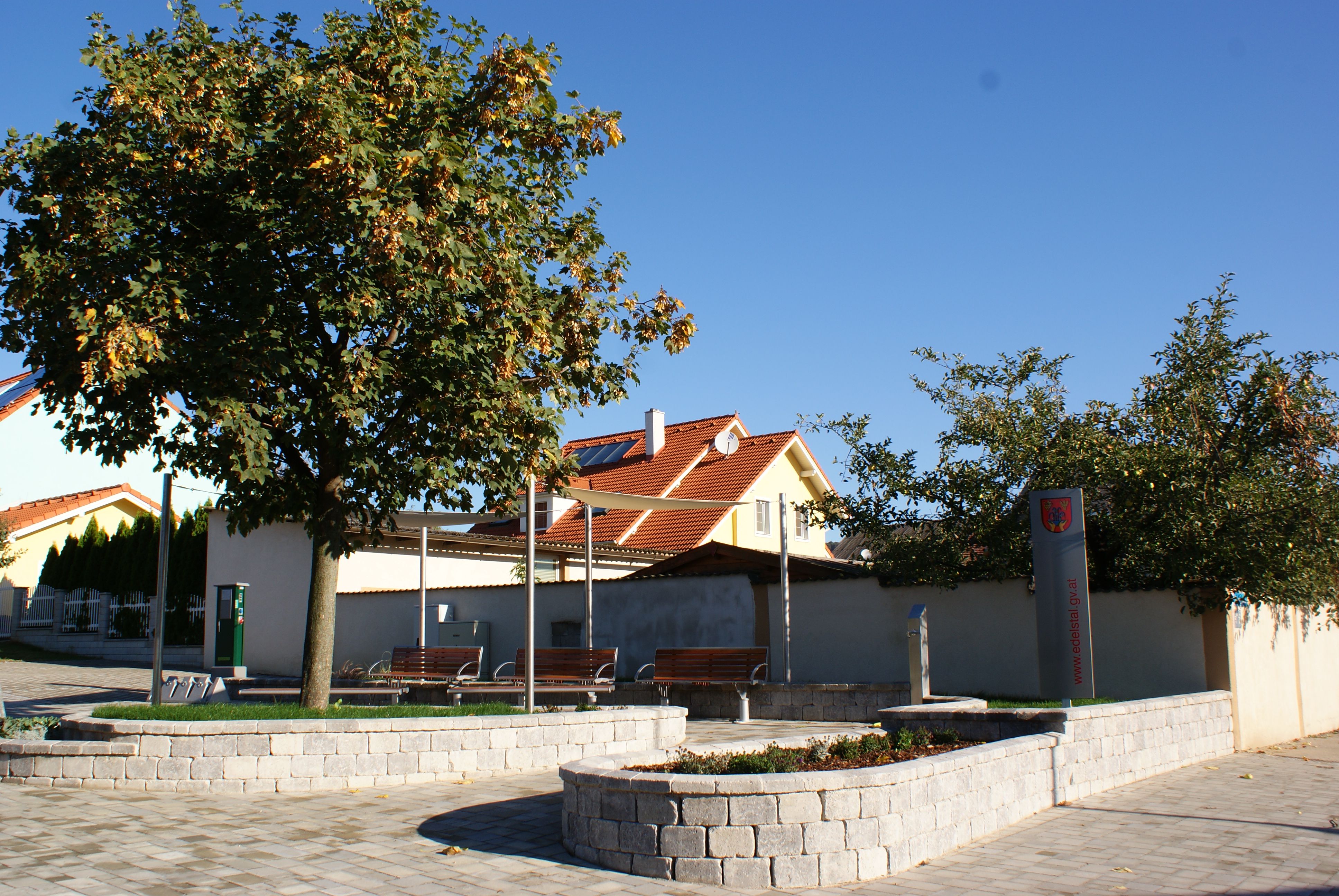Ein Radrastplatz mit Bänken, Bäumen und einem Schild vor einem Gebäude mit rotem Dach.