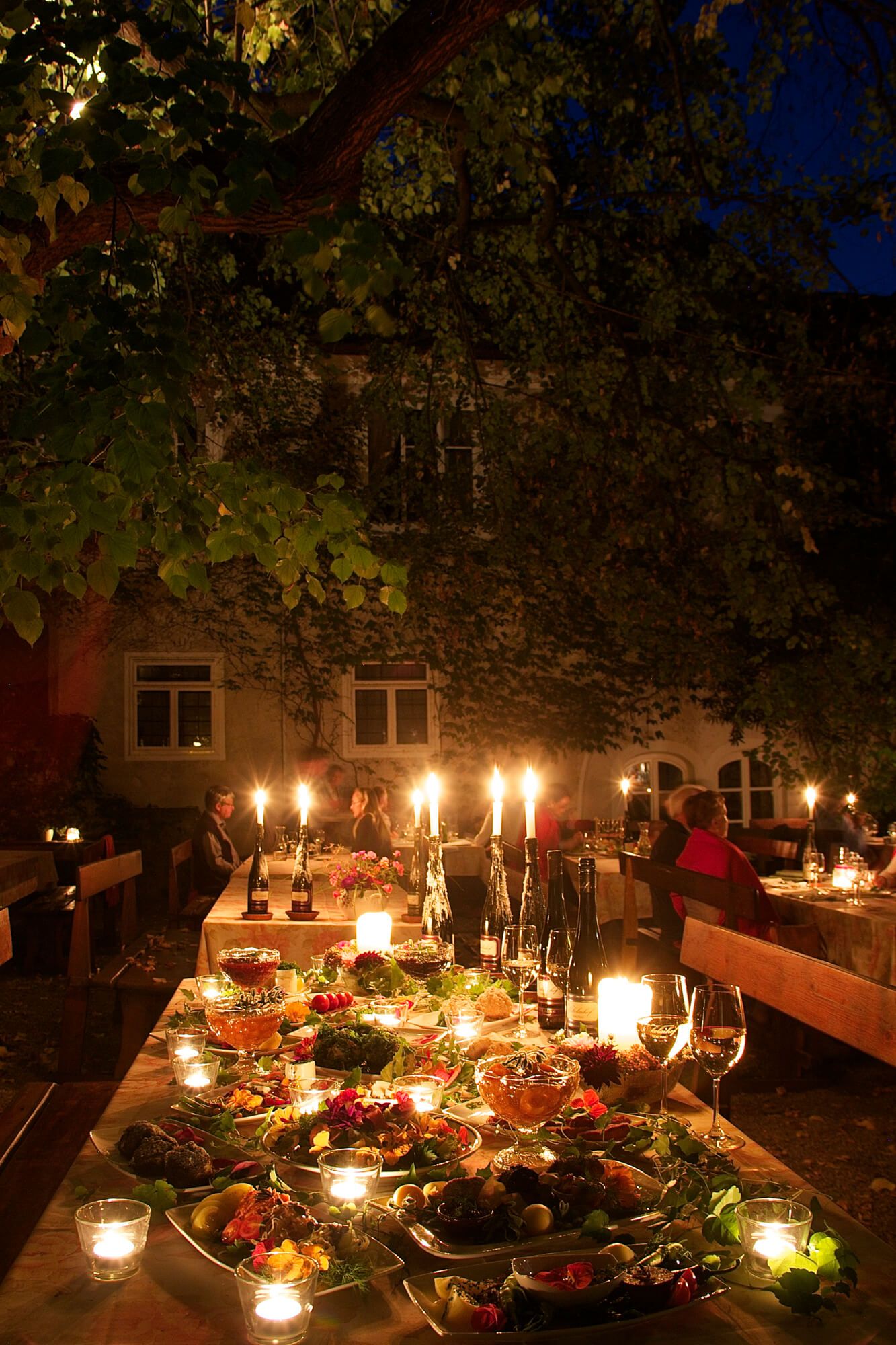 Ein festlich gedeckter Tisch im Freien mit Kerzenlicht und einem Buffet aus verschiedenen Speisen. Menschen sitzen an Tischen im Hintergrund.
