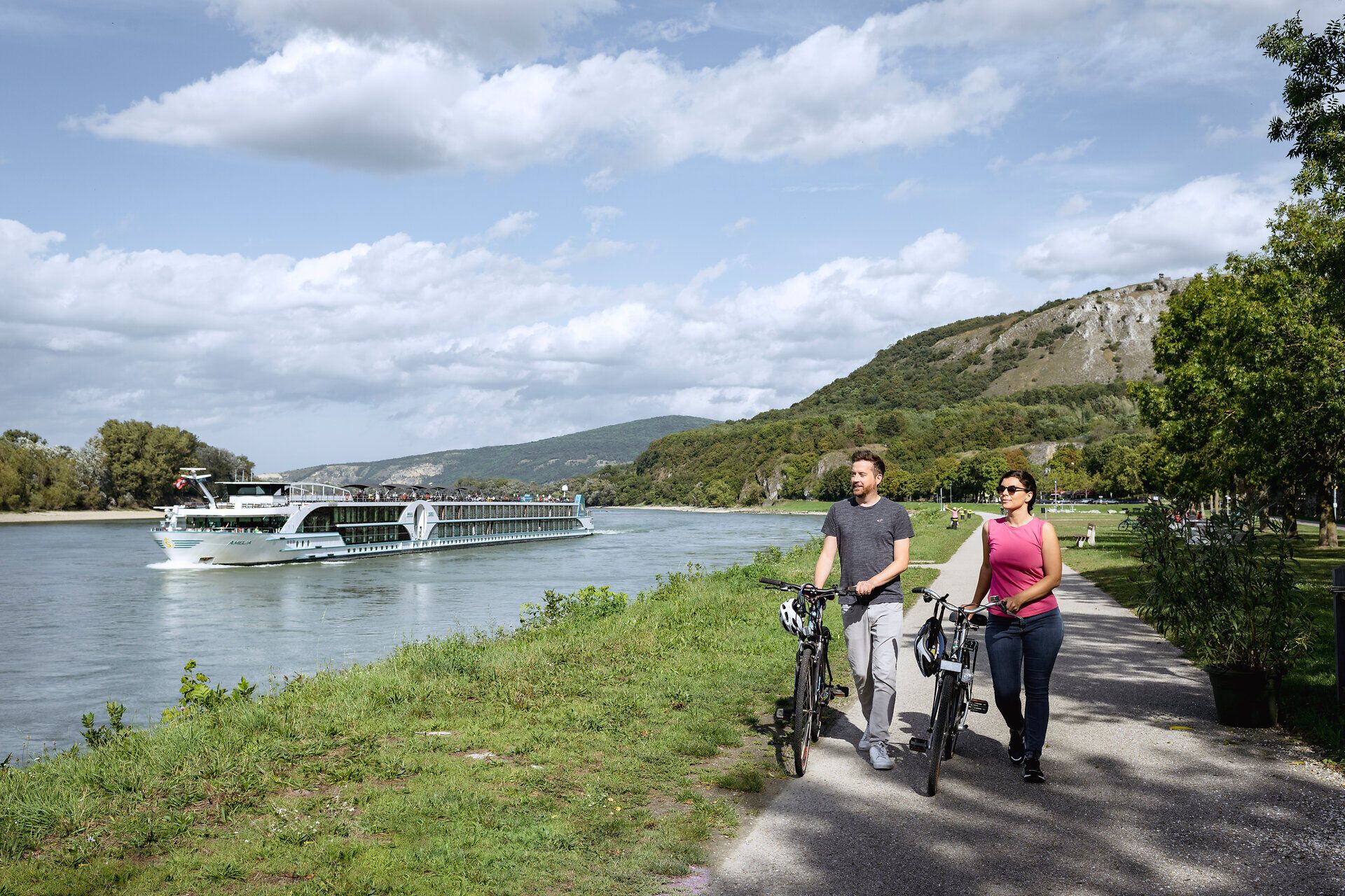 Ein Paar schiebt seine Fahrräder entlang der Donaulände, während ein großes Kabinenschiff vorbeifährt.