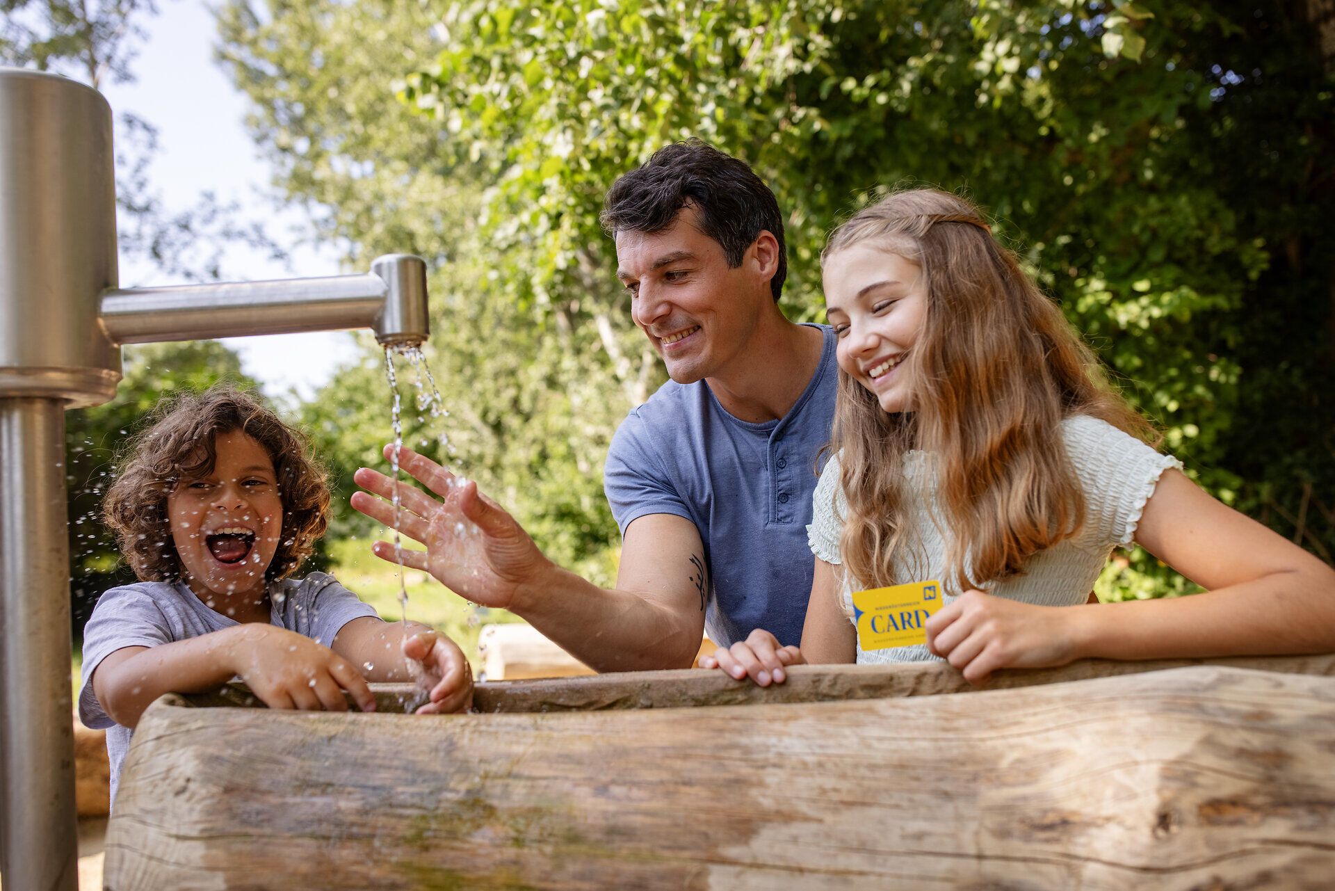 Ein fröhliches Zusammensein von Kindern und Erwachsenen am Wasser, umgeben von üppigem Grün. Das plätschernde Wasser und die strahlenden Gesichter schaffen eine einladende Atmosphäre, die zum Spielen und Entdecken einlädt.