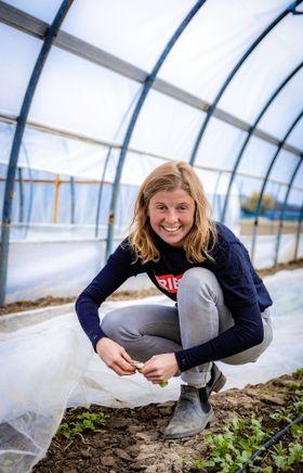 Junge Frau hockt lachend im Gewächshaus bei Gemüsebeeten.