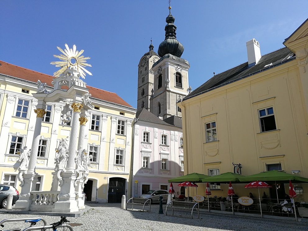 Historischer Platz mit barocker Säule, Kirche und Café.