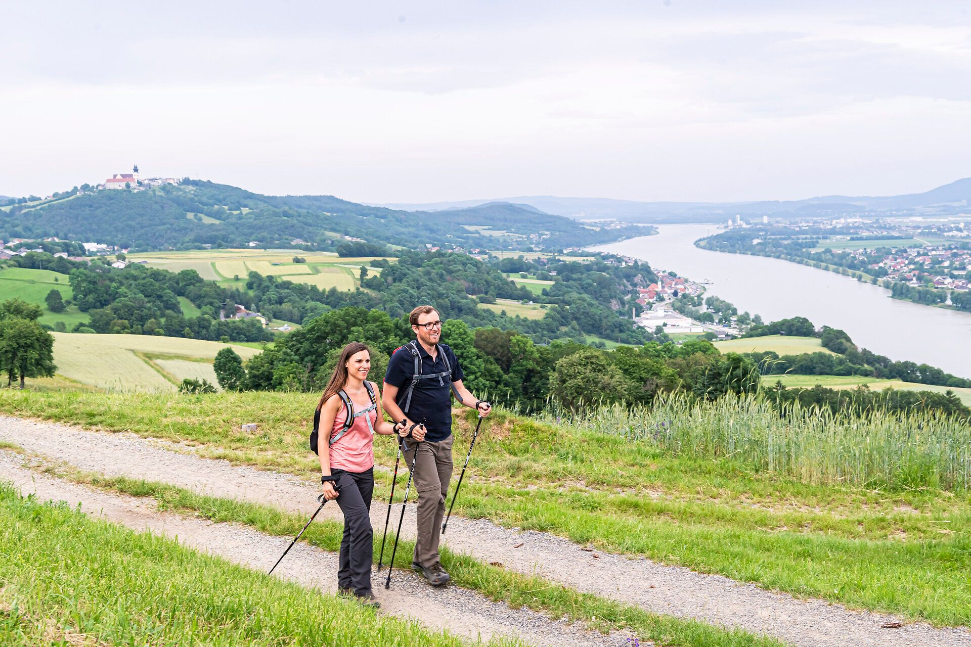 Zwei Wanderer genießen die frische Bergluft und die atemberaubende Aussicht auf die sanften Hügel und den glitzernden Fluss. Umgeben von üppigem Grün und blühenden Wildblumen, erleben sie den perfekten Sommertag in der Natur.
