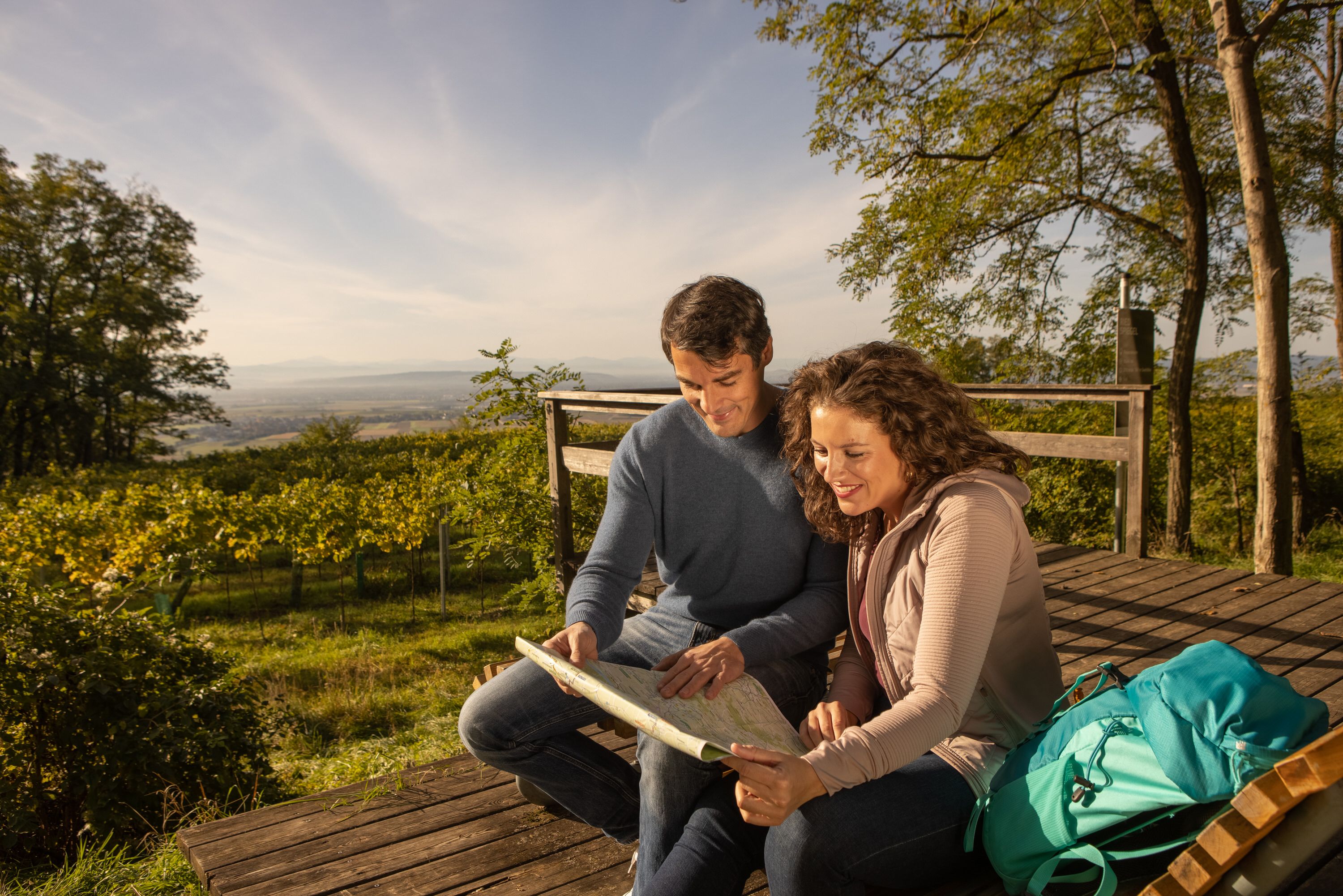 Ein Paar sitzt auf einer Holzplattform inmitten von Weinbergen, betrachtet gemeinsam eine Wanderkarte und genießt den Ausblick über die herbstliche Landschaft.