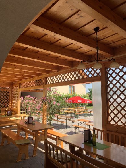 Holzpergola mit Tischen und Bänken, Blick auf einen Garten mit roten Sonnenschirmen.