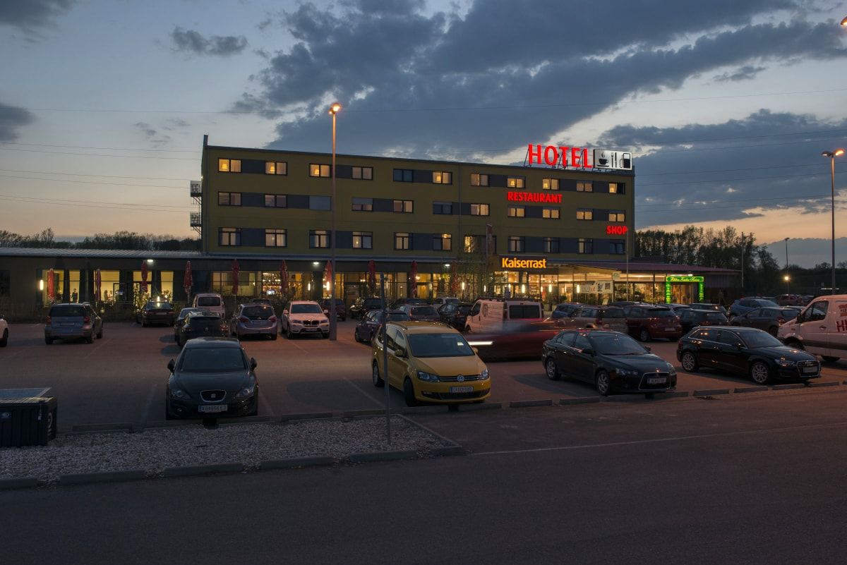 Abendaufnahme eines Hotels mit Parkplatz und Autos davor.