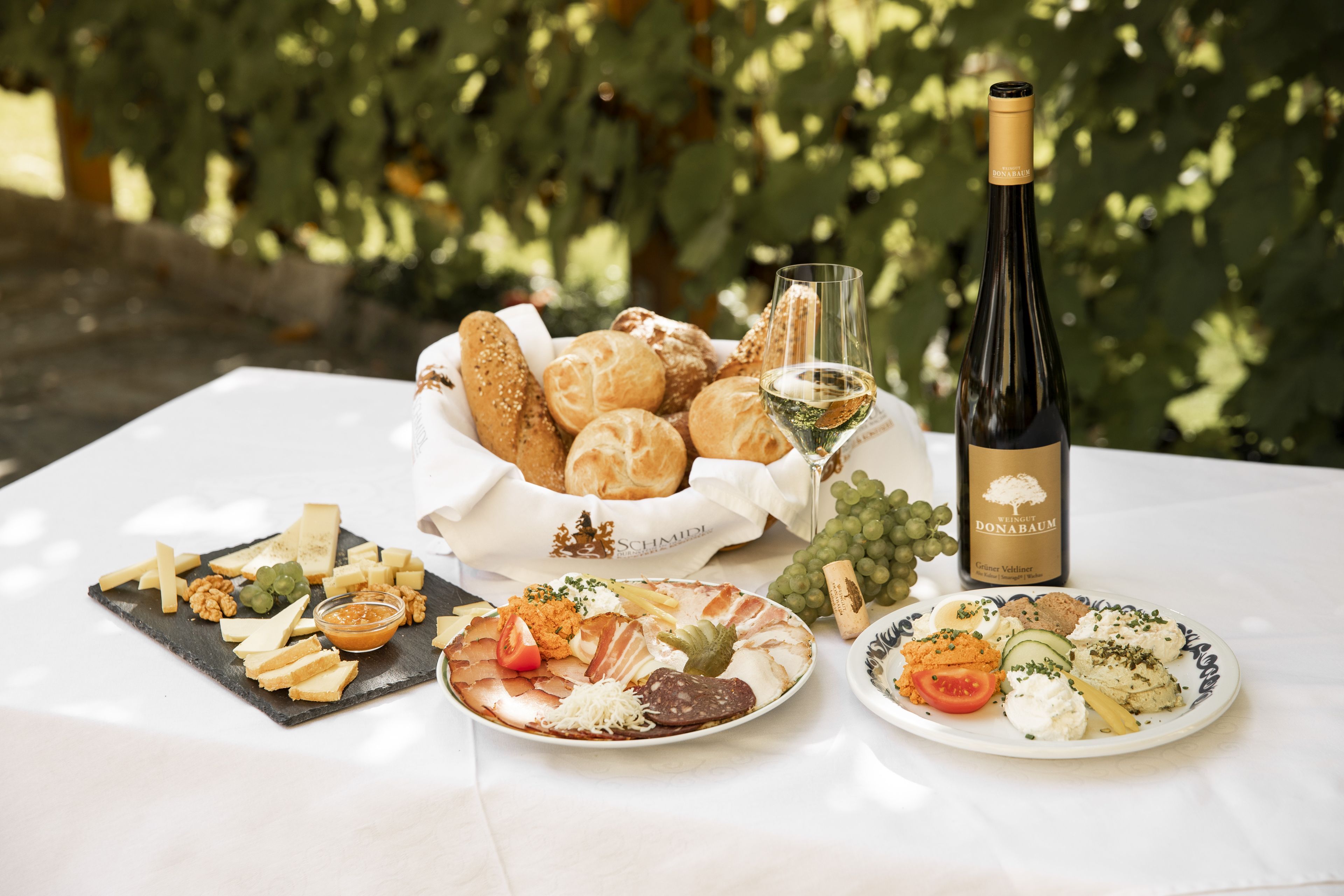 Heurigenjause mit Brot, Käse, Aufschnitt, Wein und Trauben auf einem Tisch.