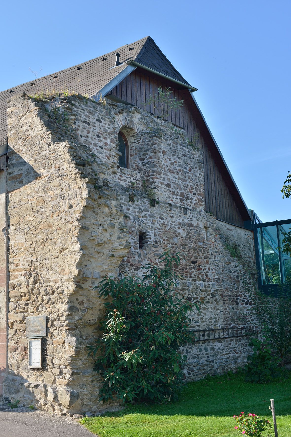 Steinmauer eines historischen Gebäudes mit modernem Anbau und Pflanzen im Vordergrund.