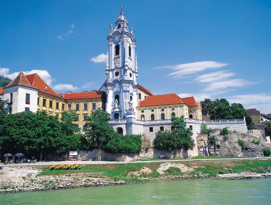Barockkirche mit blau-weißer Fassade und rotem Dach an einem Flussufer.