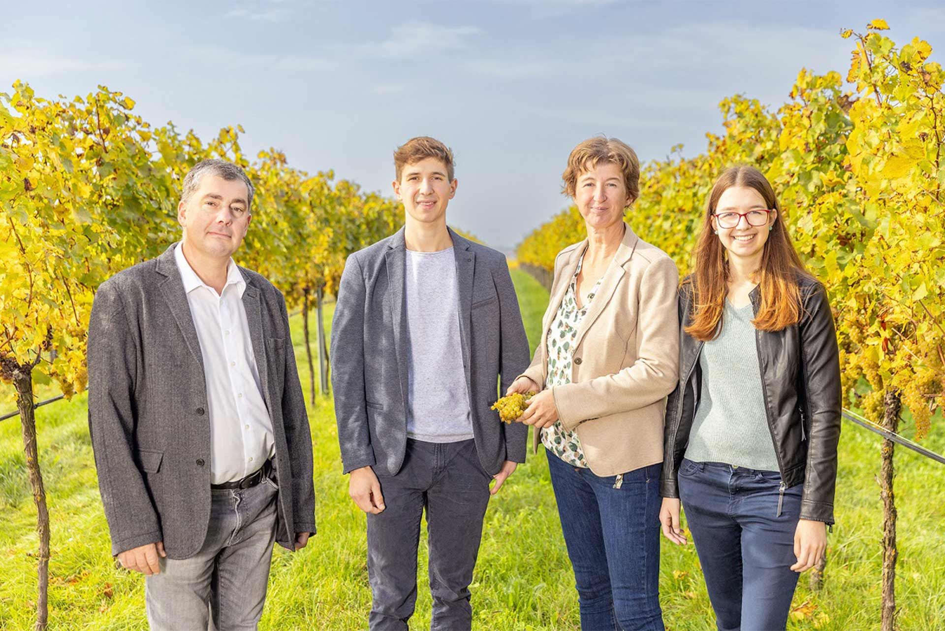 Vier Personen stehen gemeinsam im Weingarten zwischen den Rebreihen; natürliche, entspannte Atmosphäre mit ländlichem Hintergrund.