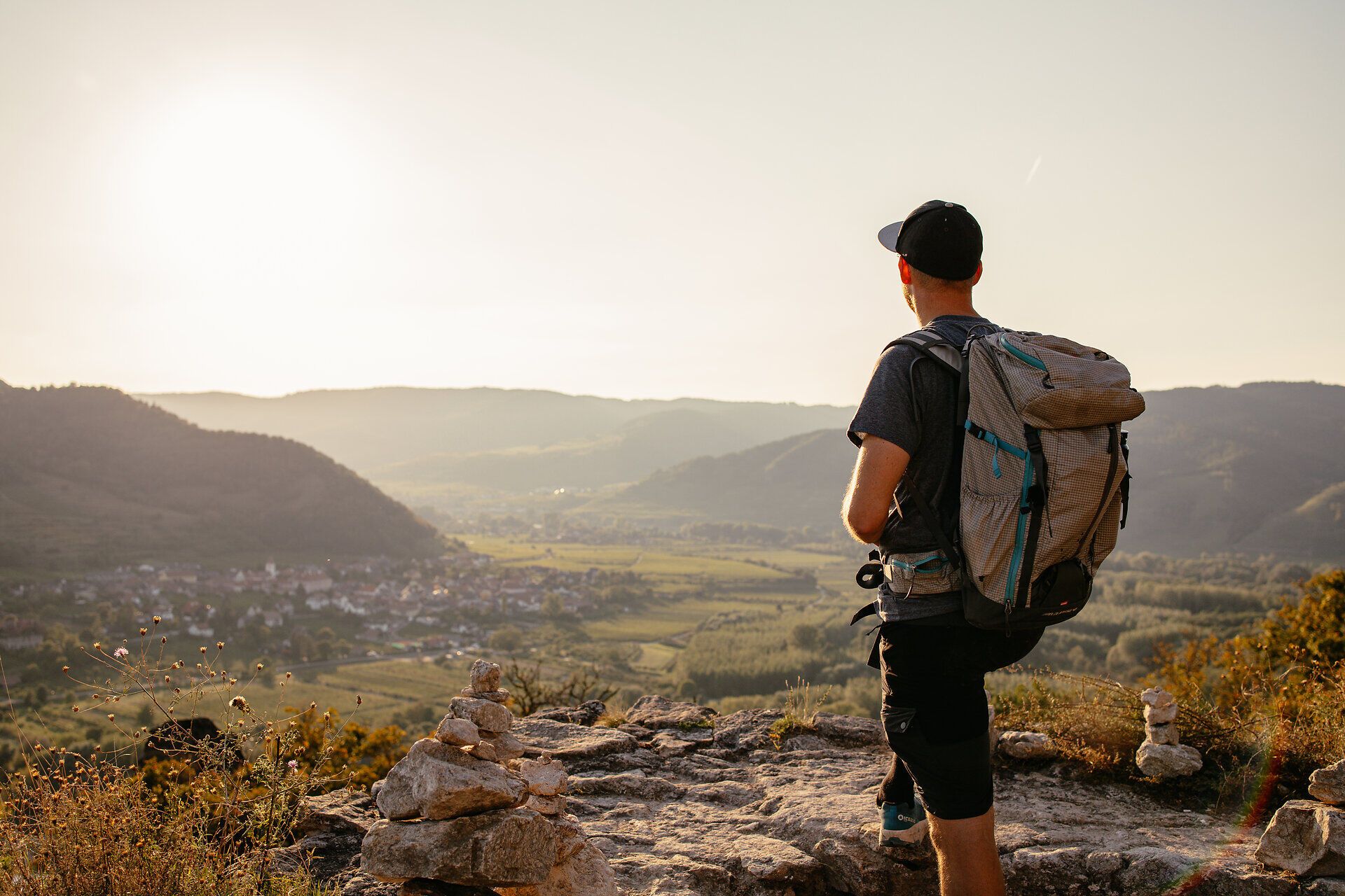 Ein Wanderer genießt den atemberaubenden Blick über die sanften Hügel und das malerische Tal, während die Sonne langsam am Horizont untergeht. Die goldenen Strahlen tauchen die Landschaft in ein warmes Licht und laden dazu ein, die Schönheit der Natur zu erleben.