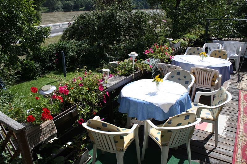 Terrasse mit Tischen und Stühlen, umgeben von Pflanzen und Blumen, mit Blick auf einen Fluss.