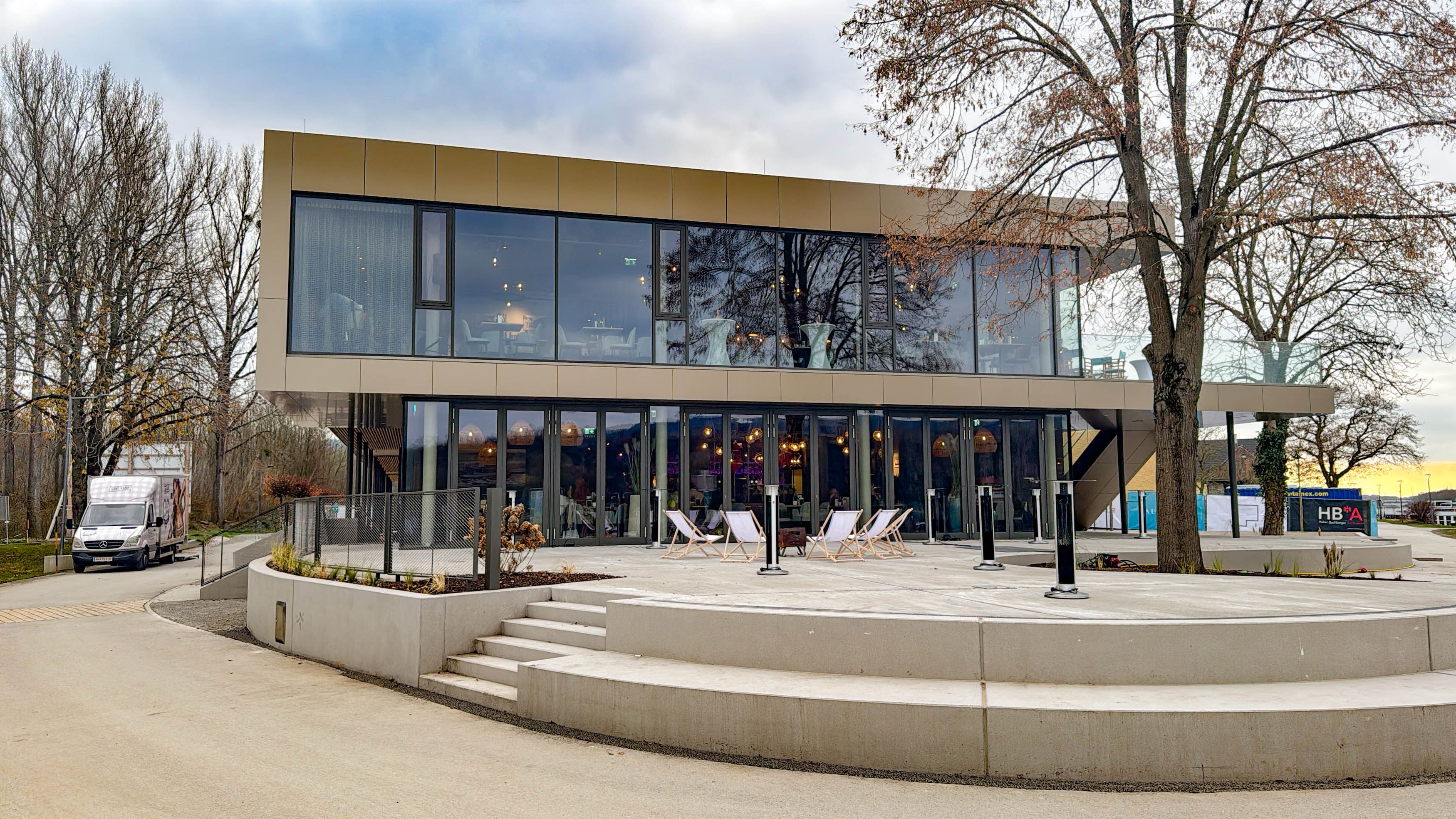 Moderne zweistöckige Gebäude mit Glasfassade und Terrasse, umgeben von Bäumen.
