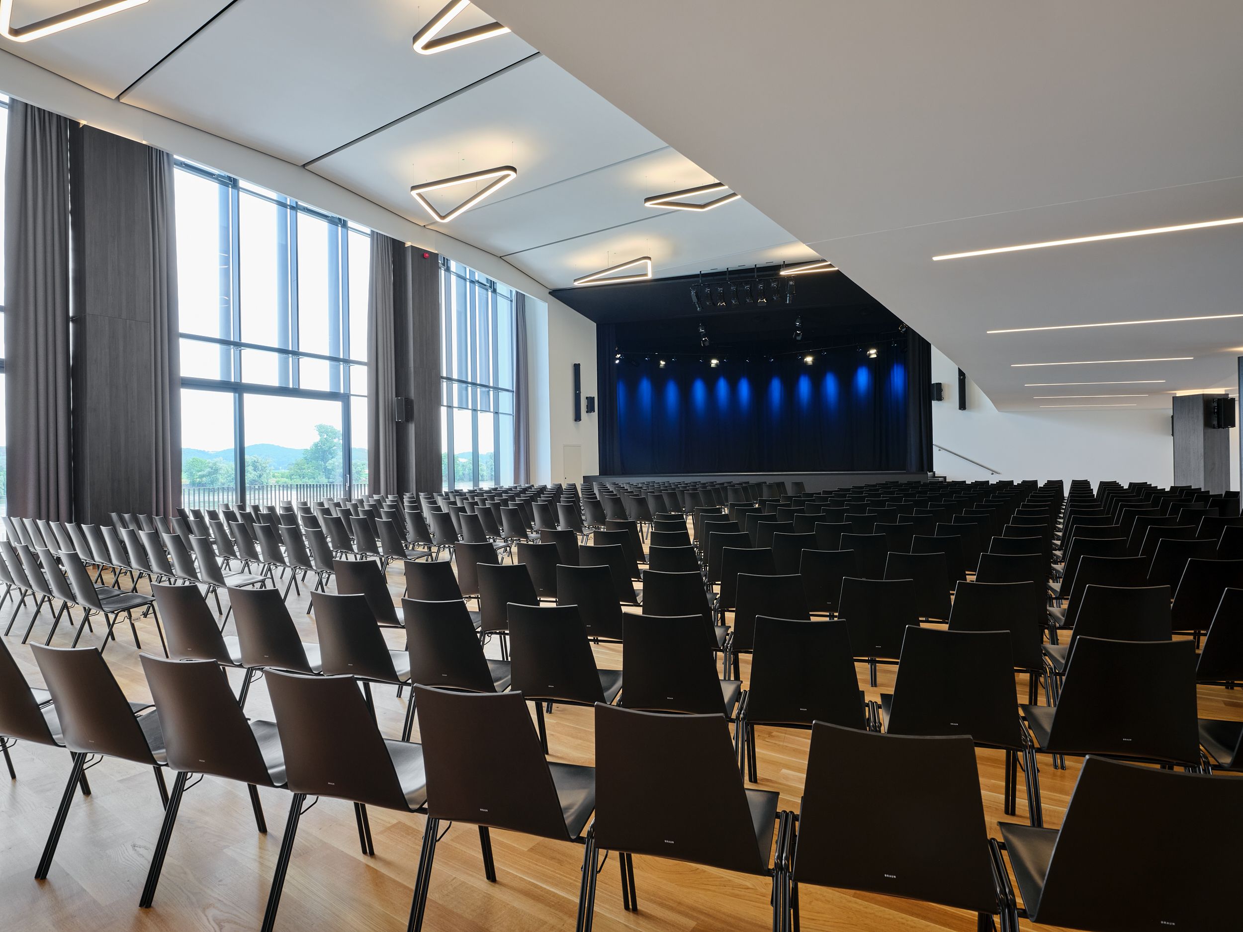 Interior view of a modern event hall with empty black chairs and a stage with blue lighting.