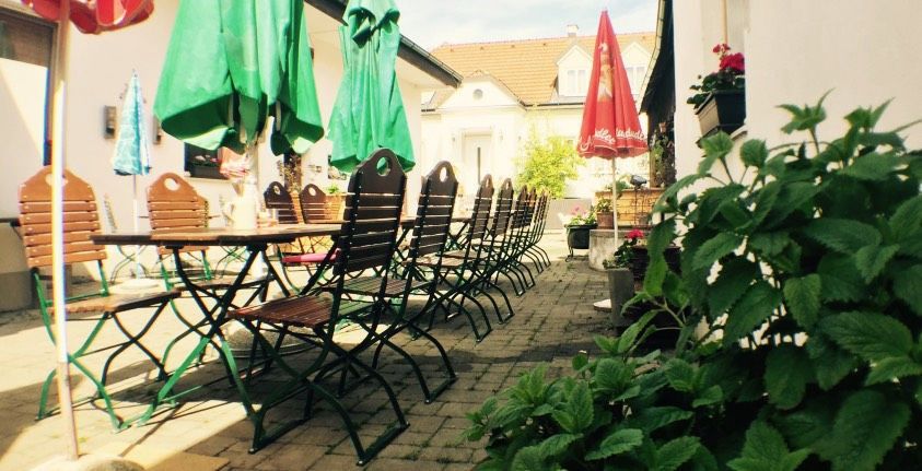 Ein leerer Gastgarten mit Holztischen und -stühlen, umgeben von grünen Sonnenschirmen und Pflanzen.