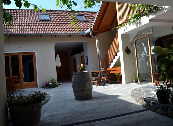 Ein gemütlicher Innenhof mit Holzmöbeln, Pflanzen und einem Weinfass. Ein Gebäude mit roten Dachziegeln und Holztreppe im Hintergrund.