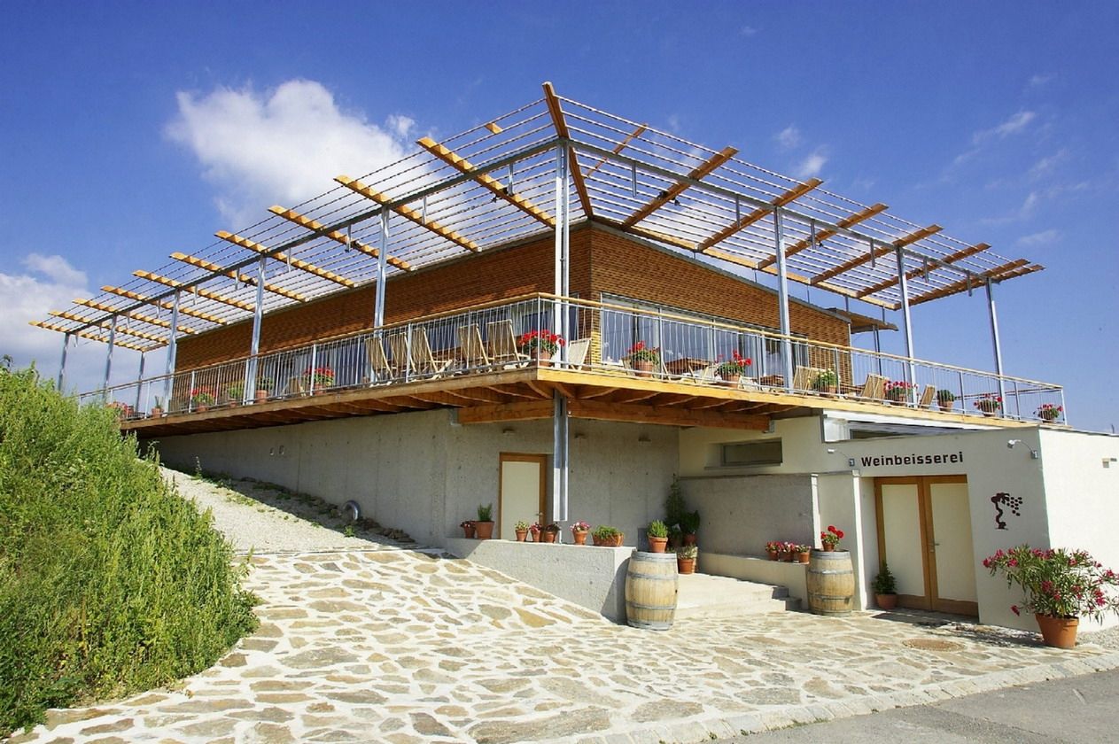 Modernes Gebäude mit Terrasse und Pergola, umgeben von Pflanzen und Weinfässern, unter blauem Himmel.