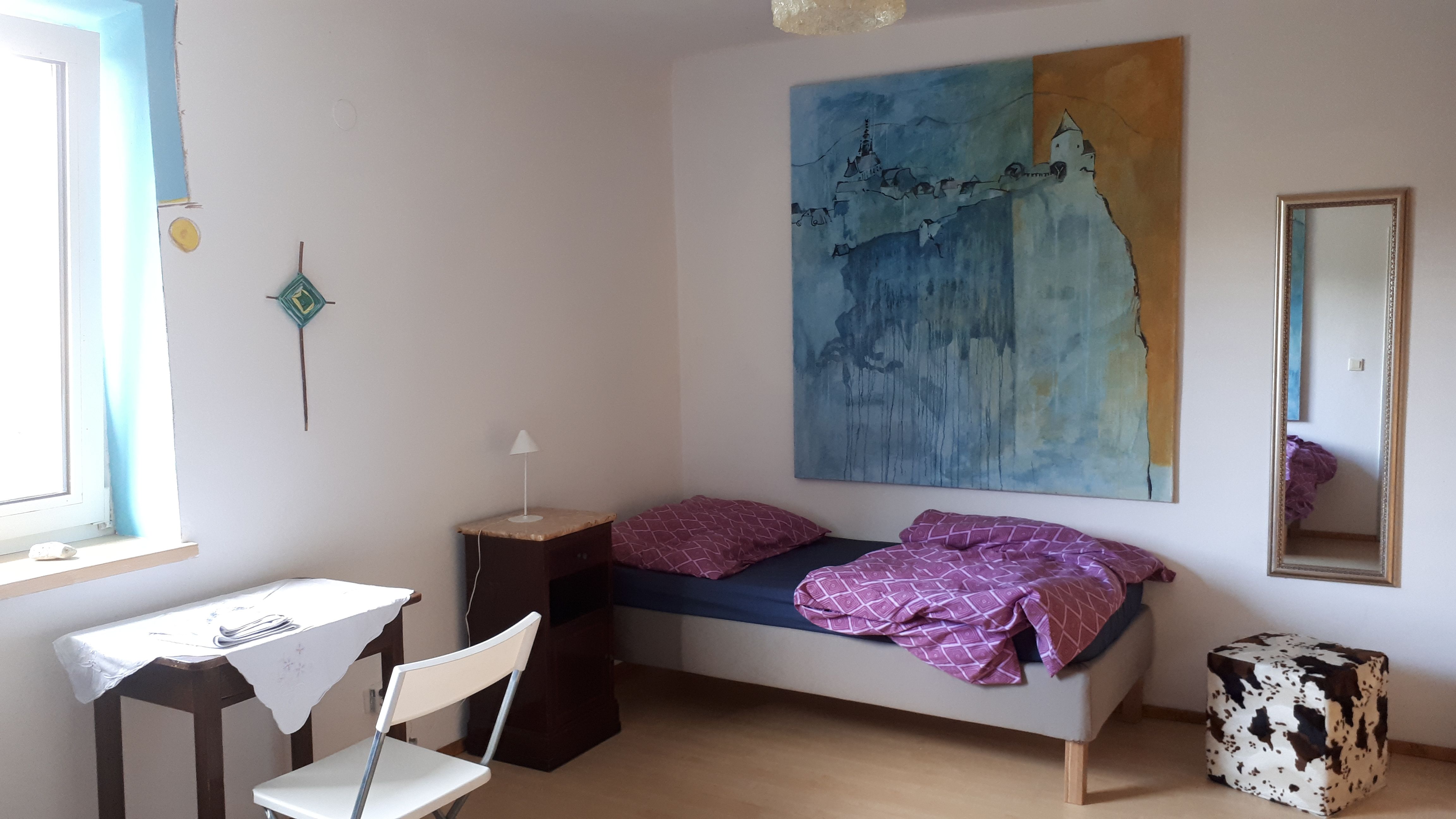 Ein einfach eingerichtetes Zimmer mit Bett, Tisch, Stuhl und großem Gemälde an der Wand.