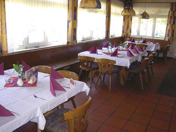 Ein Gastraum mit gedeckten Tischen, weißen Tischdecken und lila Servietten. Holzstühle und große Fenster sind sichtbar.