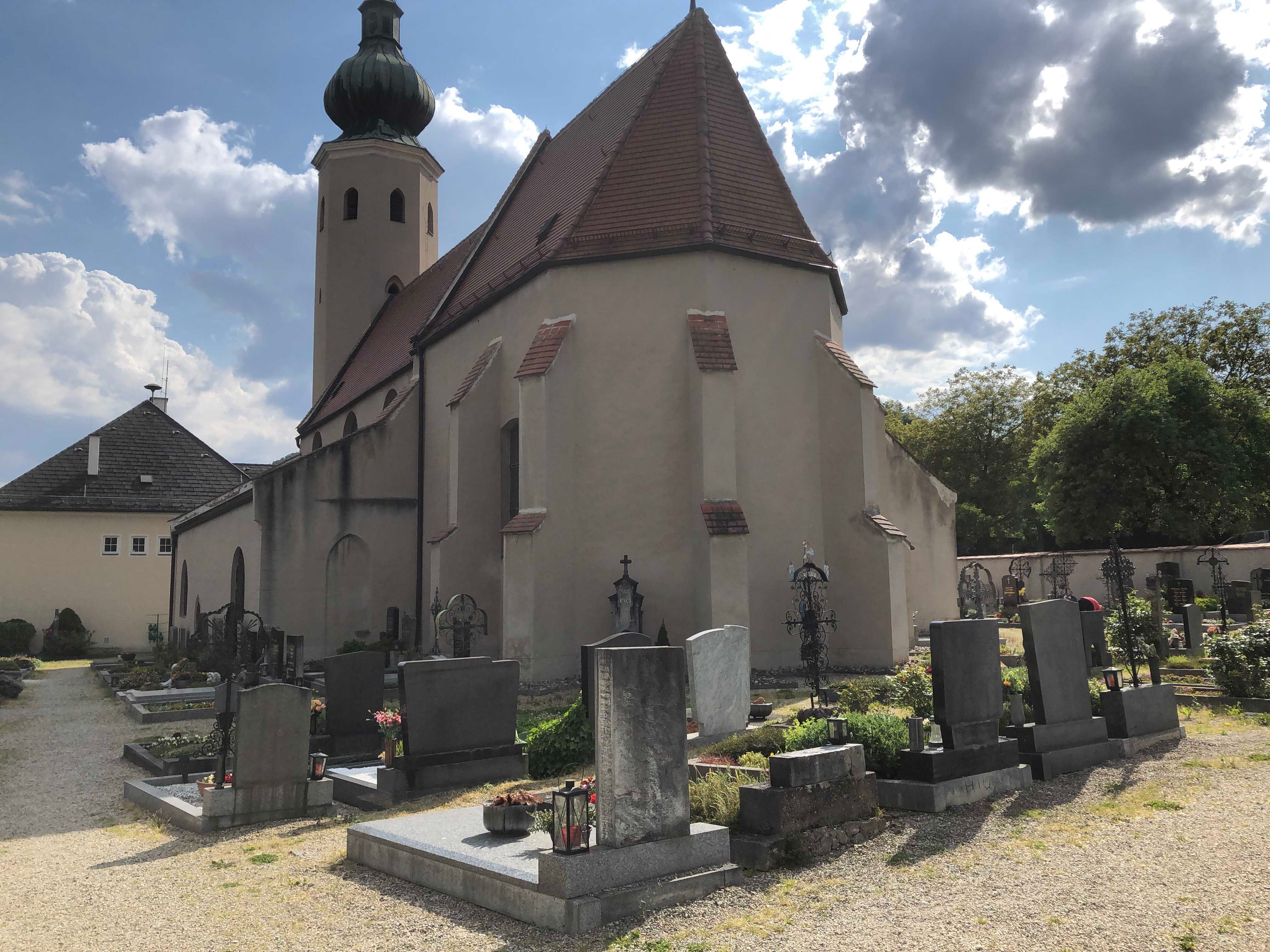 Templom temetővel és sírkövekkel az előtérben, felhős égbolt.