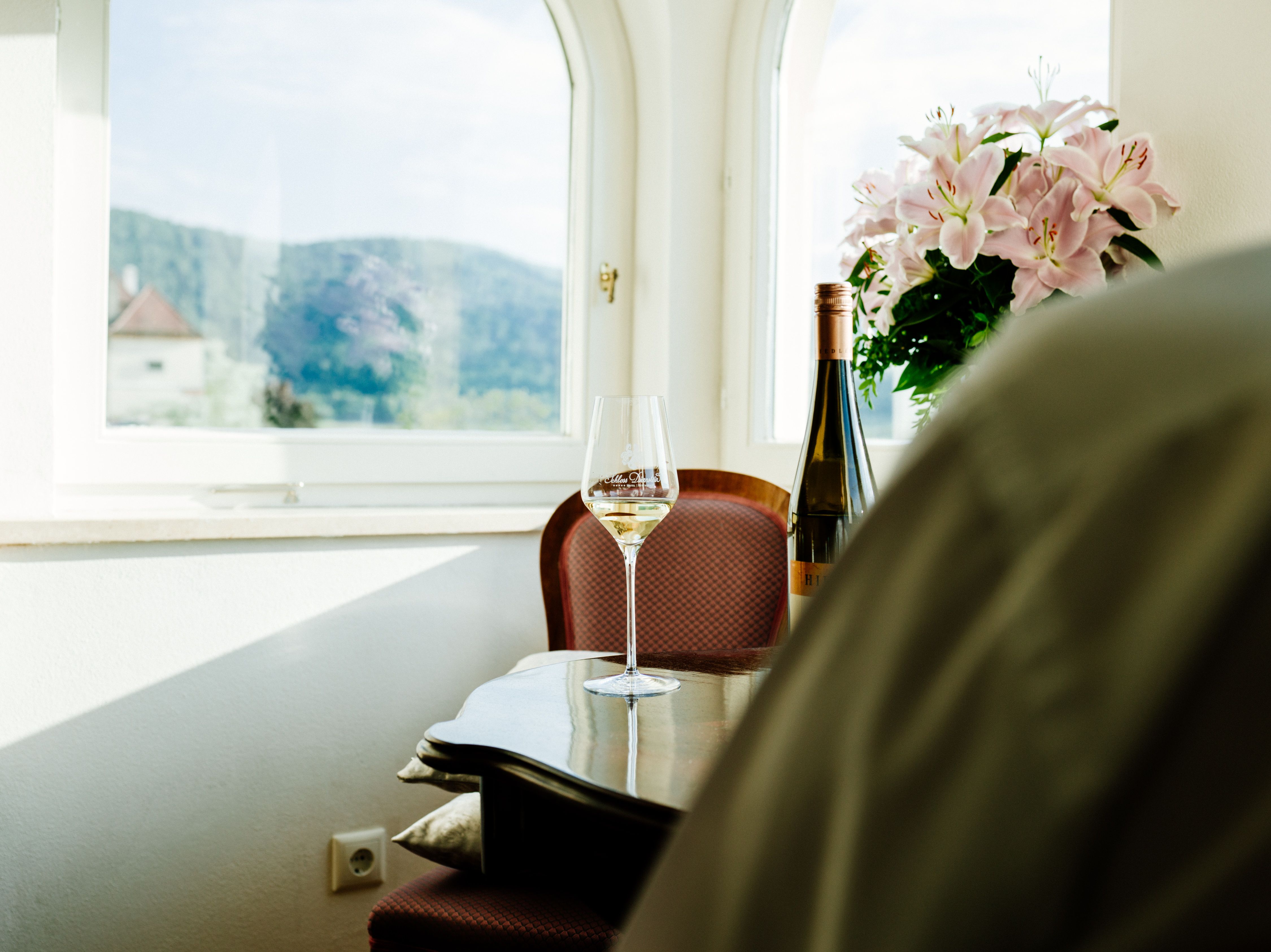 Ein Weinglas und eine Weinflasche auf einem Tisch vor einem Fenster mit Blick auf Hügel und Blumen.