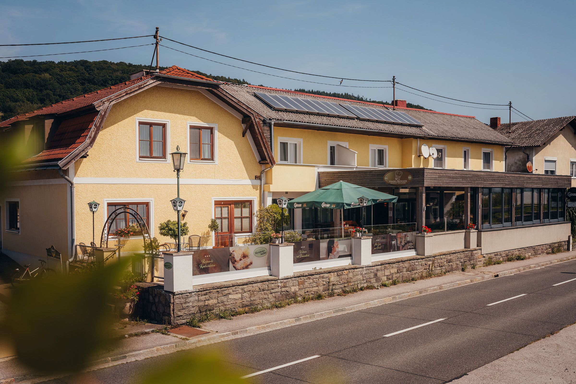 Gelbes Gasthaus mit Terrasse und Sonnenschirmen an einer Landstraße.