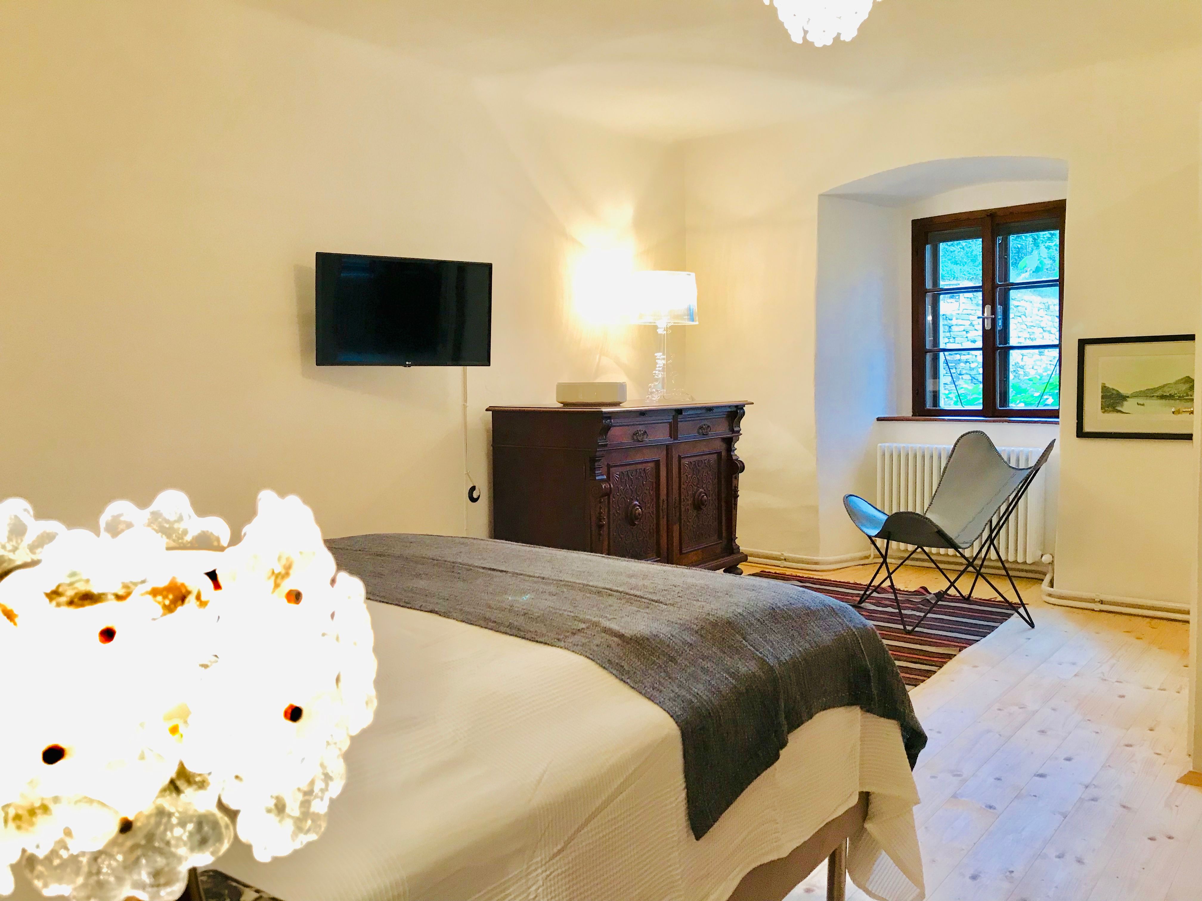 Cozy bedroom with bed, antique chest of drawers, armchair and wall-mounted TV.
