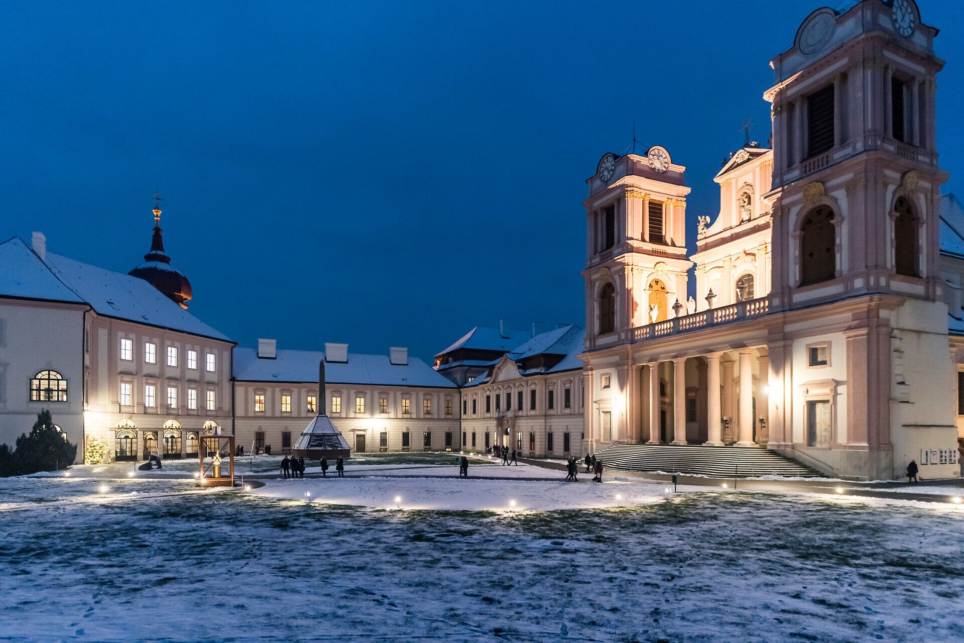 In der winterlichen Dämmerung erstrahlt die barocke Architektur in warmem Licht und schafft eine einladende Atmosphäre. Der sanfte Schnee bedeckt den Platz und verleiht der Szenerie eine magische Note, während Besucher den Adventmarkt erkunden und die festliche Stimmung genießen.