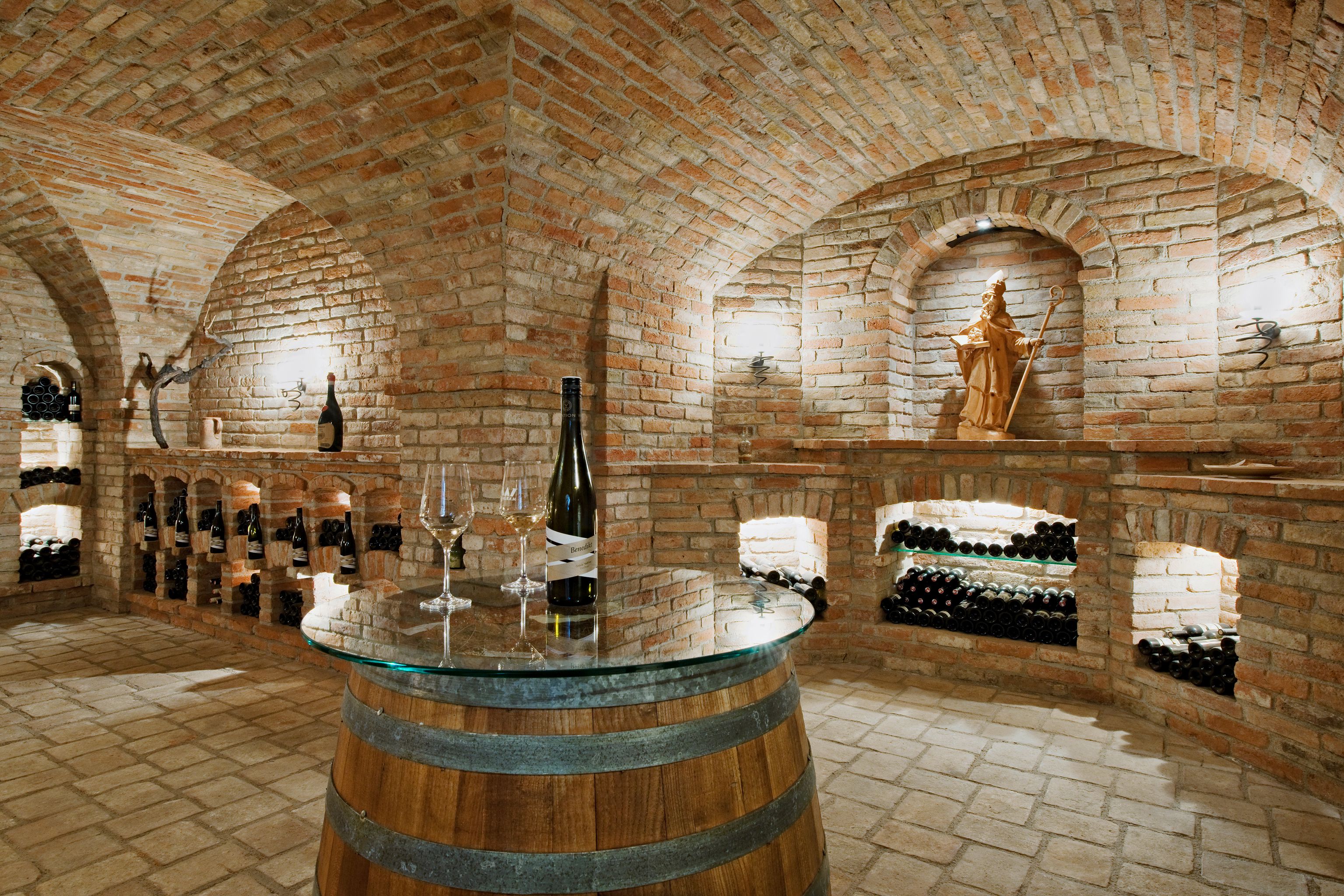 Ein Gewölbekeller mit Ziegelwänden, Weinflaschen in Regalen und einem Fass mit Glasplatte als Tisch.