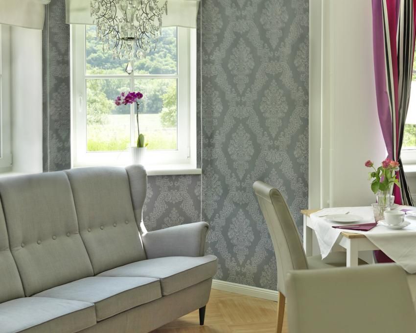 Innenraum eines stilvollen Wohnzimmers mit grauem Sofa, dekorativer Tapete, Kronleuchter und Esstisch mit Blumen.