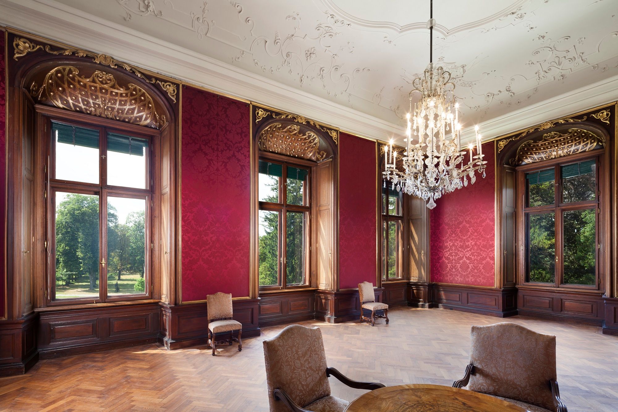 Eleganter Raum im Schloss Eckartsau mit roten Wänden, großen Fenstern und einem Kronleuchter.