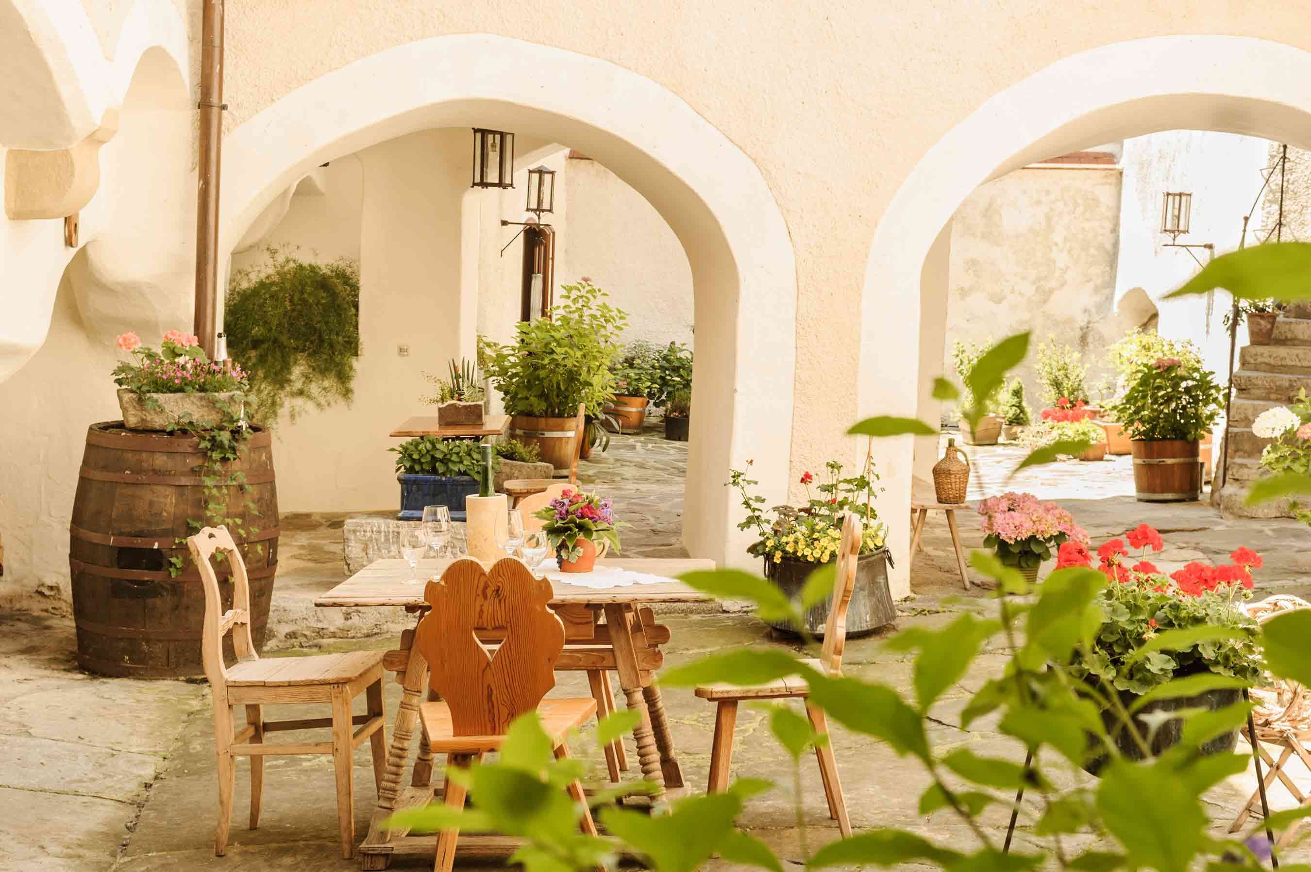 Ein gemütlicher Innenhof mit Holztisch und Stühlen, umgeben von Pflanzen und Blumen in Töpfen.