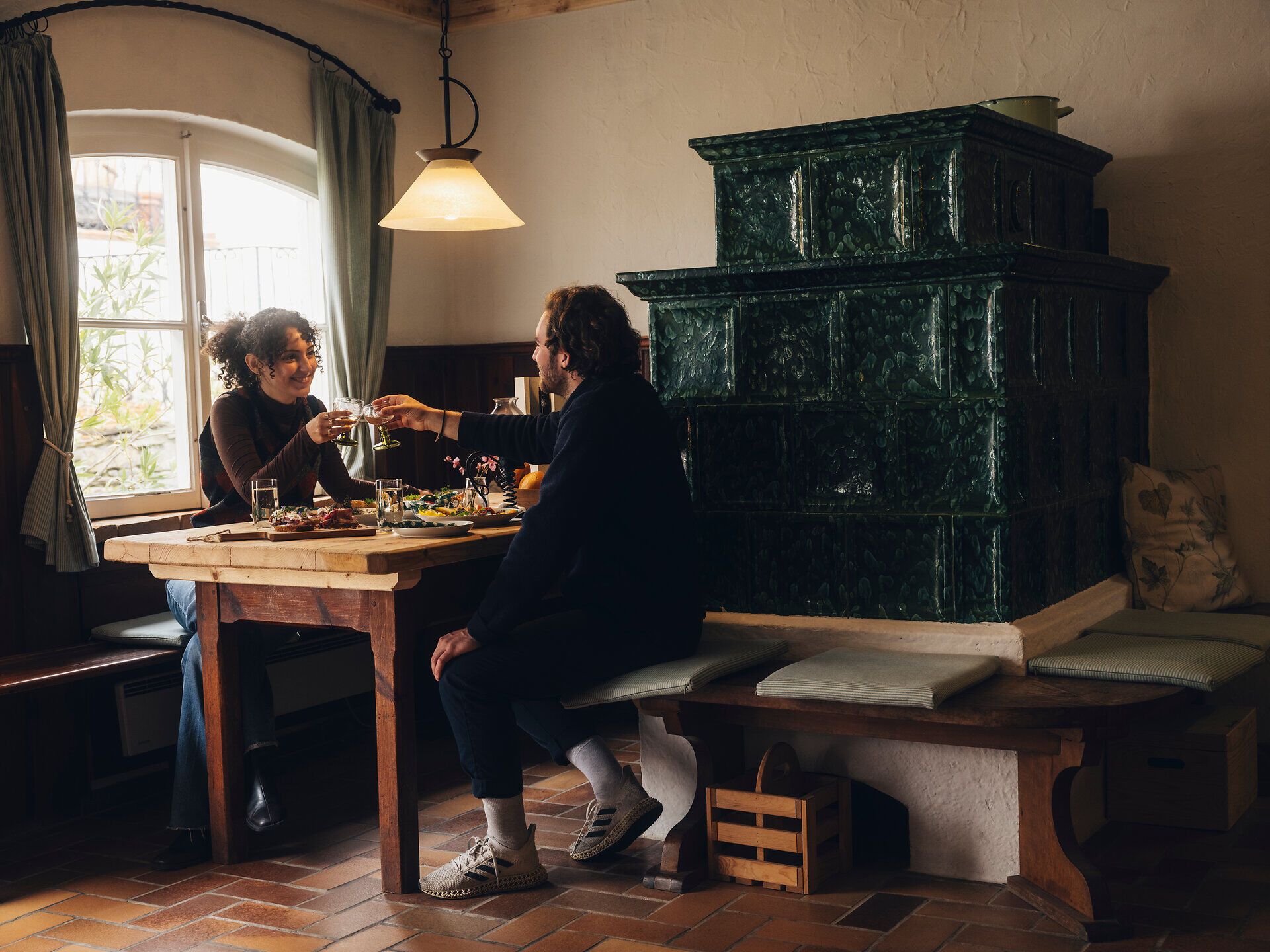 Zwei Personen sitzen an einem Holztisch in einer gemütlichen Stube und stoßen mit Gläsern an; neben ihnen steht ein großer grüner Kachelofen, warmes Licht fällt durch ein Fenster.