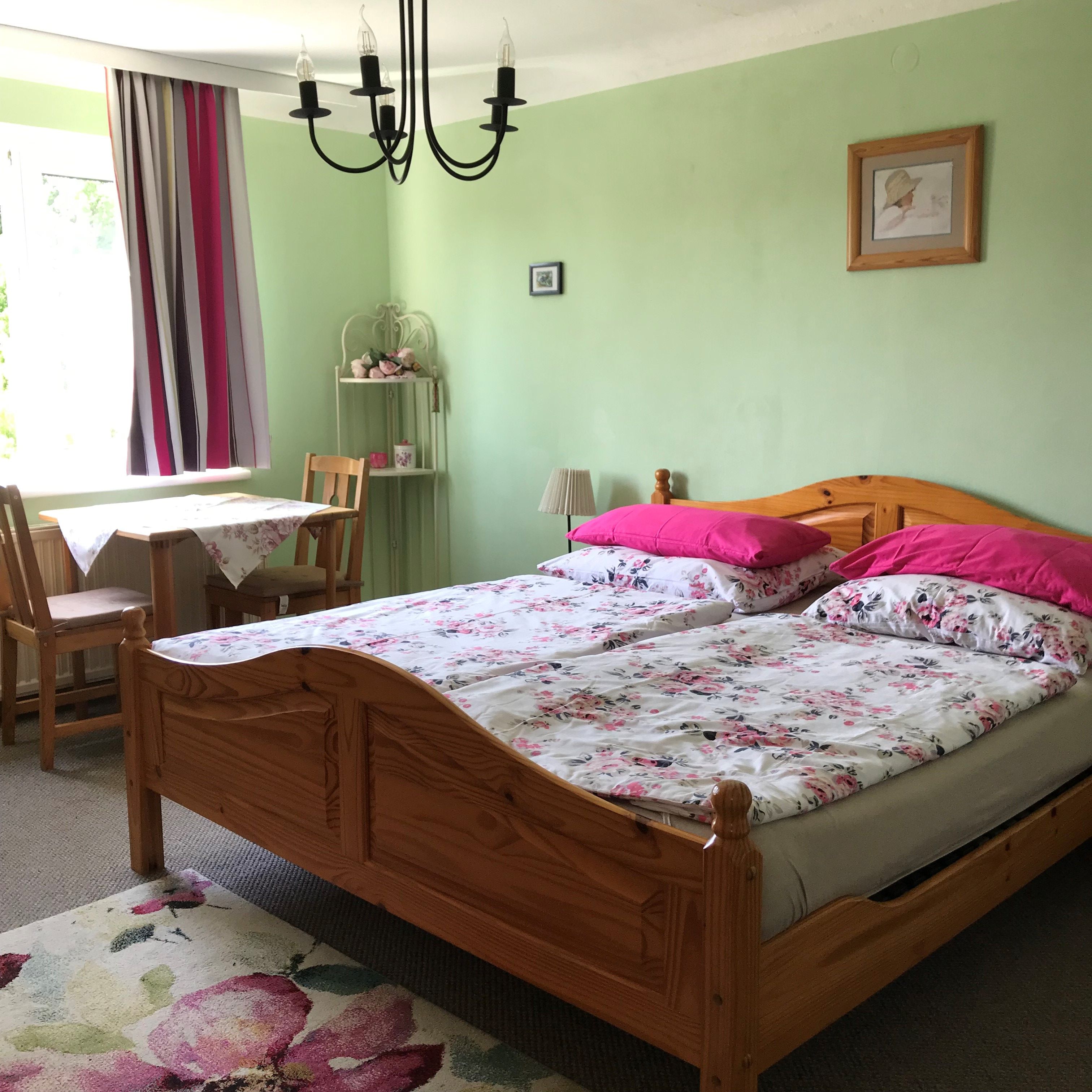 Schlafzimmer mit Doppelbett, grüne Wände, Blumenbettwäsche, Tisch mit Stühlen, Vorhänge und Kronleuchter.