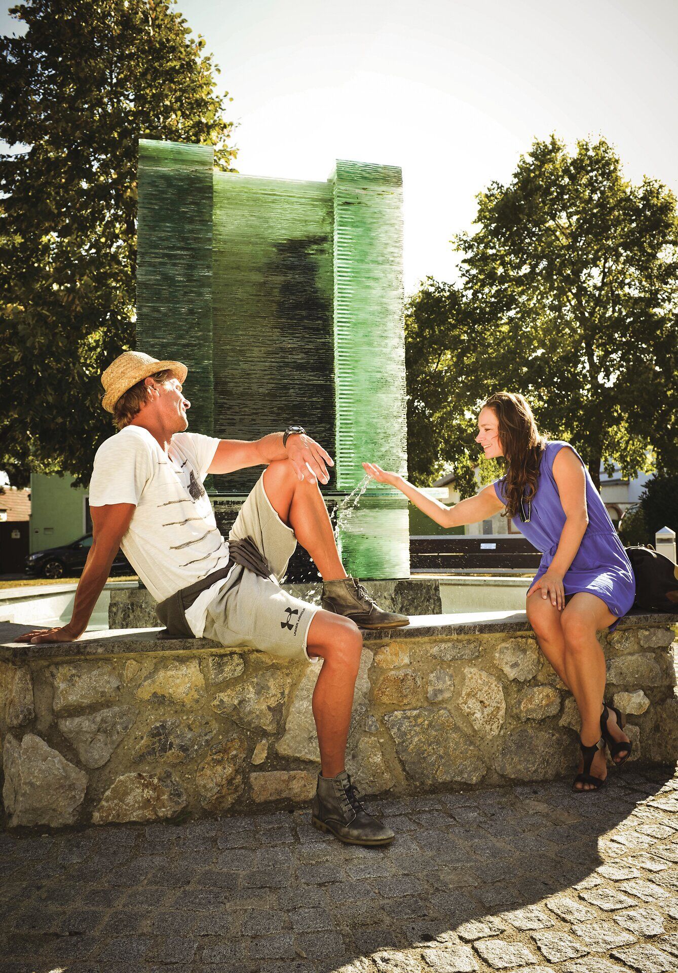 Ein Paar sitzt auf der Mauer vor einem Brunnen. Die Frau spielt mit dem Wasser. 