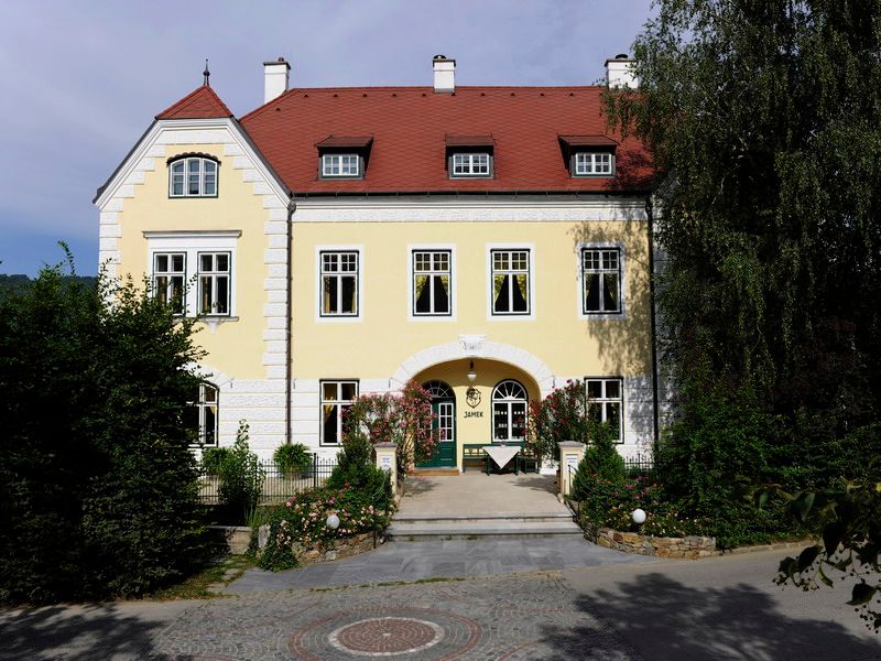 Gelbes Gebäude mit rotem Dach und grüner Tür, umgeben von Bäumen und Pflanzen.