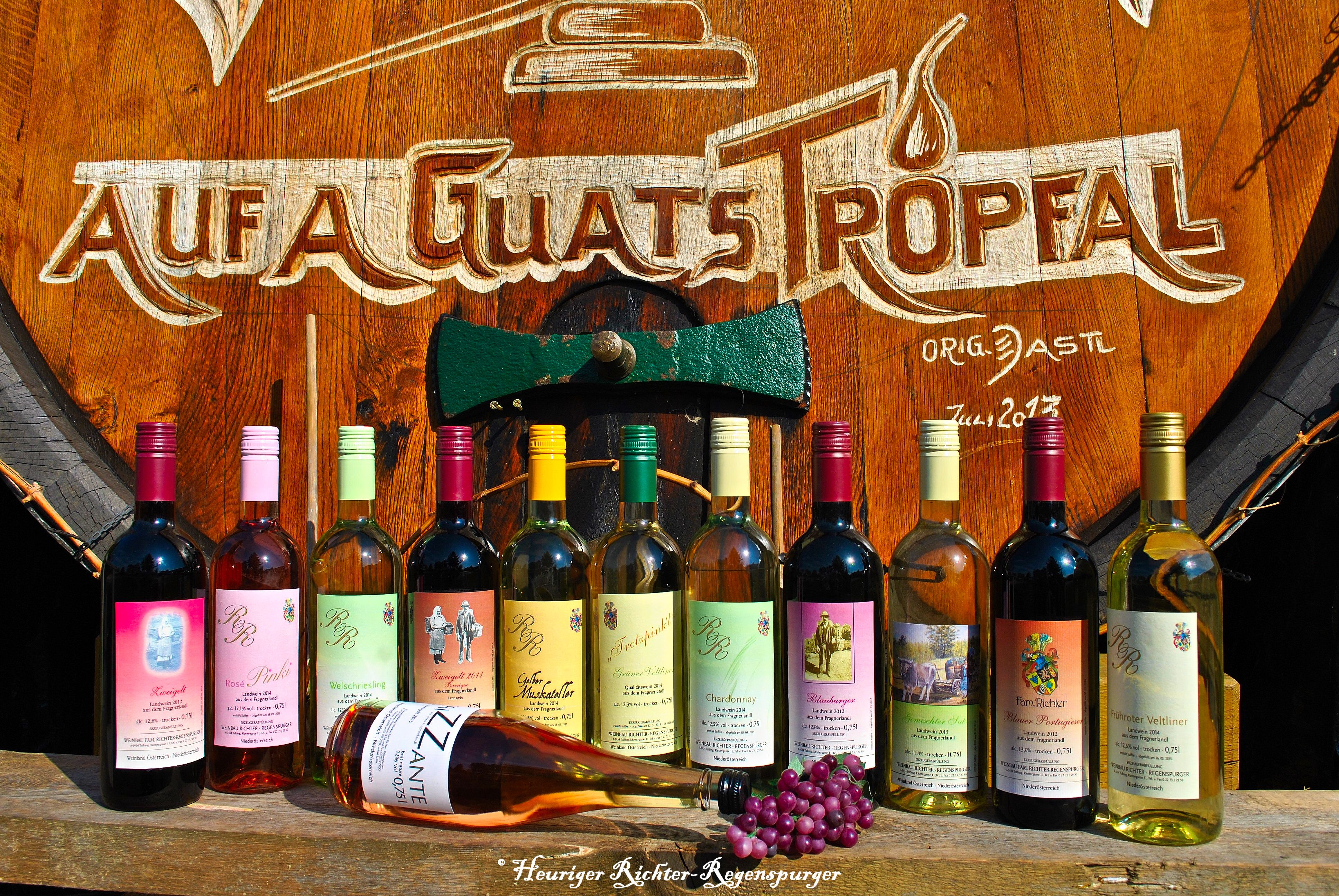 Verschiedene Weinflaschen vor einem großen Holzfass mit der Aufschrift 'Auf a guats Tropfal'.