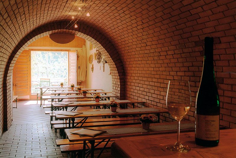 Weinkeller mit Ziegelgewölbe, Holztischen und Weinflasche.