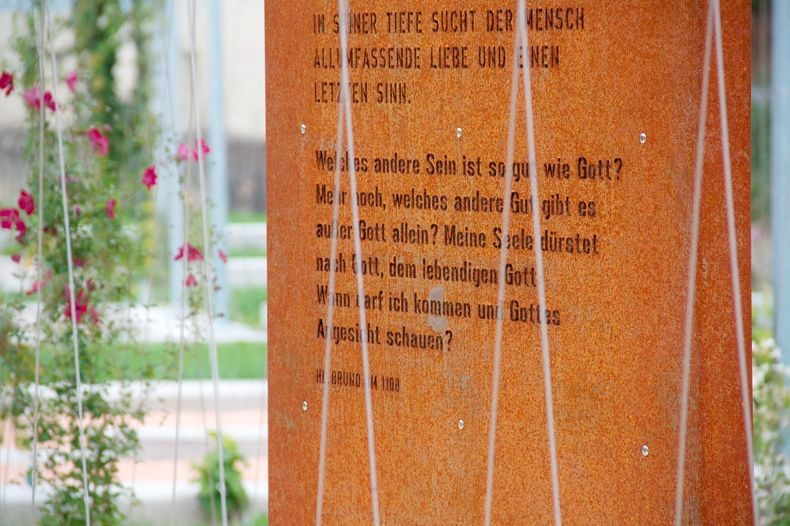 Nahaufnahme einer rostigen Metalltafel mit eingraviertem Text, umgeben von Pflanzen.