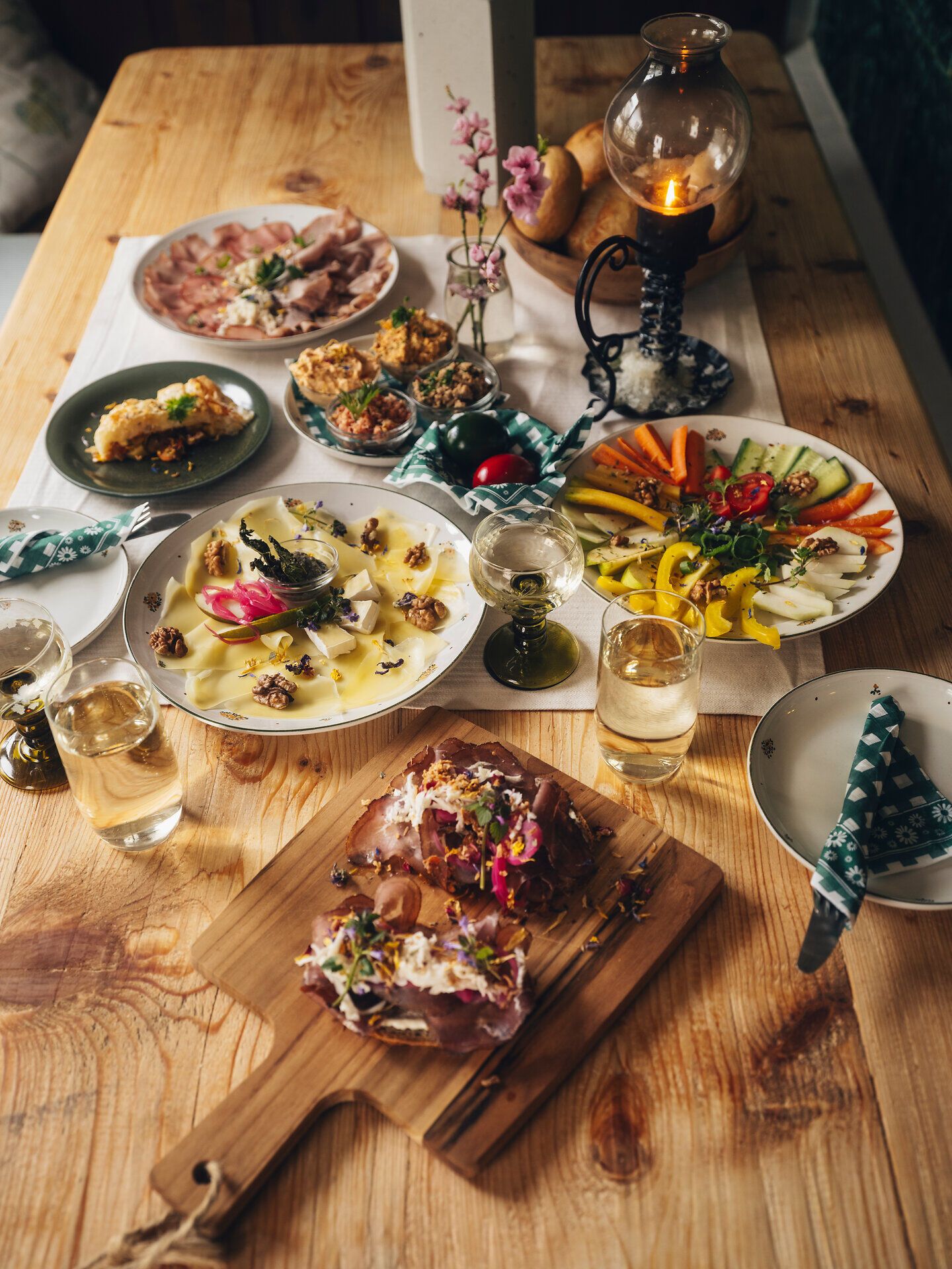 Draufsicht auf einen Holztisch mit mehreren Tellern regionaler Speisen, Brot, Gläsern und kleinen Blumenarrangements in einer gemütlichen Stube.