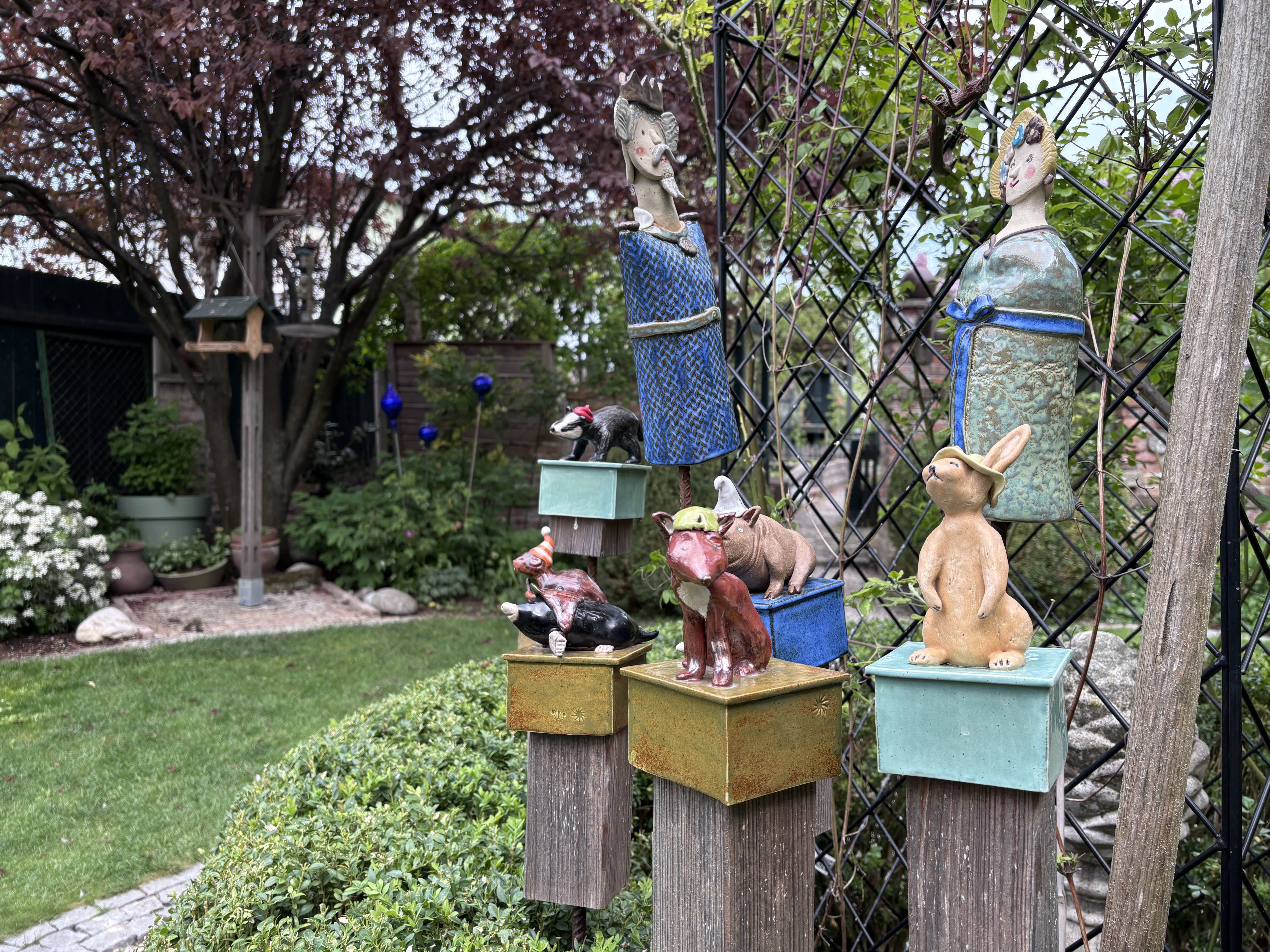 Gartenkunst mit Keramikfiguren auf Holzsäulen, umgeben von Pflanzen und einem Baum.