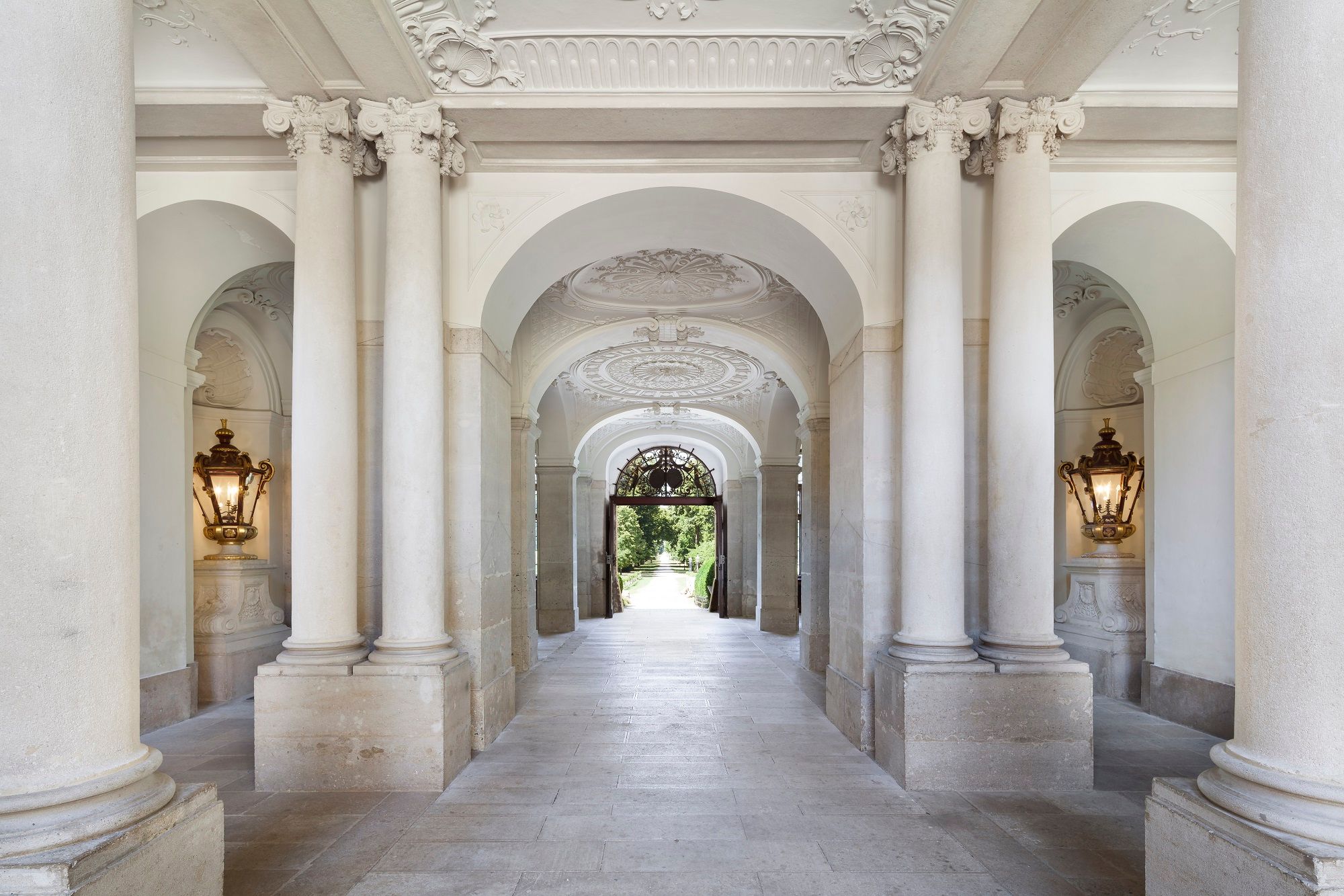Eleganter, mit Säulen gesäumter Durchgang im Schloss Eckartsau mit verzierten Decken und Wandleuchten.
