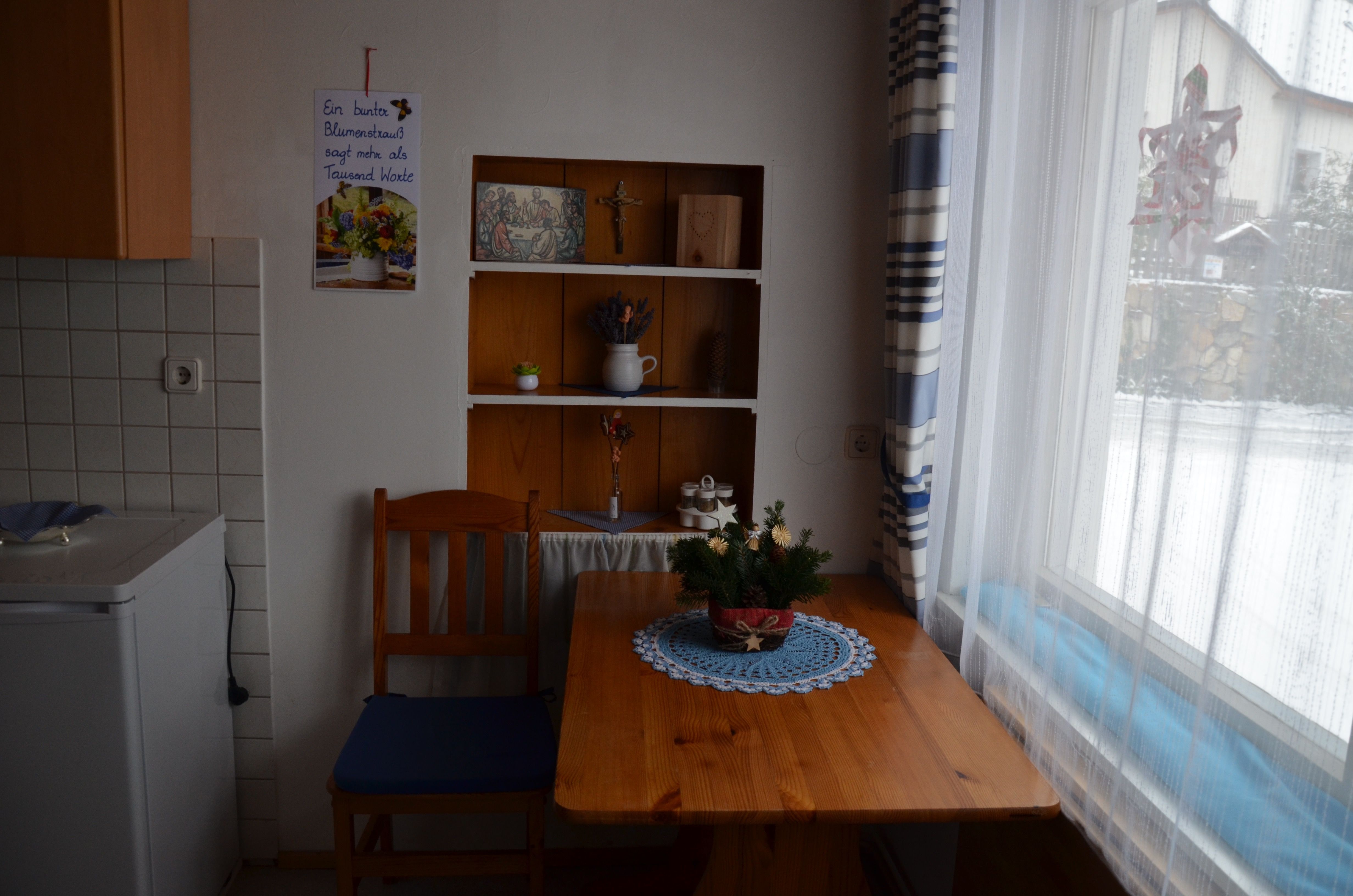 Kleine Küche mit Holztisch, Stuhl und Regal. Fenster mit Vorhang, draußen Schnee. Dekorationen und Pflanzen auf Tisch und Regal.