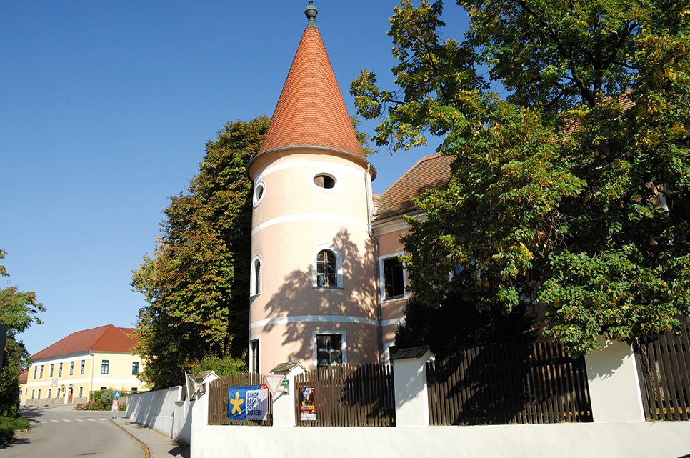 Ein rosa Gebäude mit Turm und rotem Dach in Fels am Wagram, umgeben von Bäumen und einem blauen Himmel.