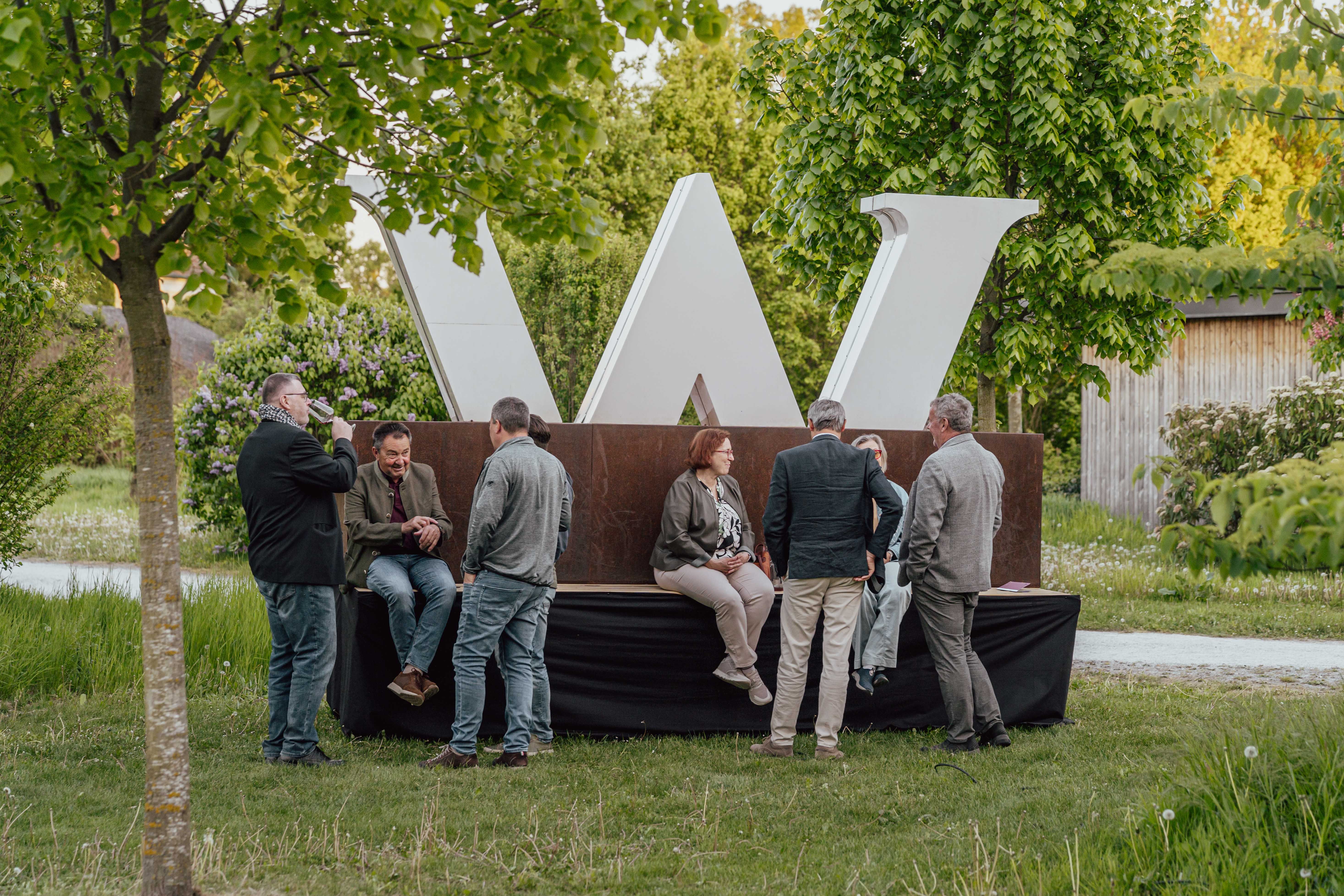 Gäste unterhalten sich bei einer Weinveranstaltung im Freien vor einer großen Wagram Skulptur in einer grünen Gartenanlage.
