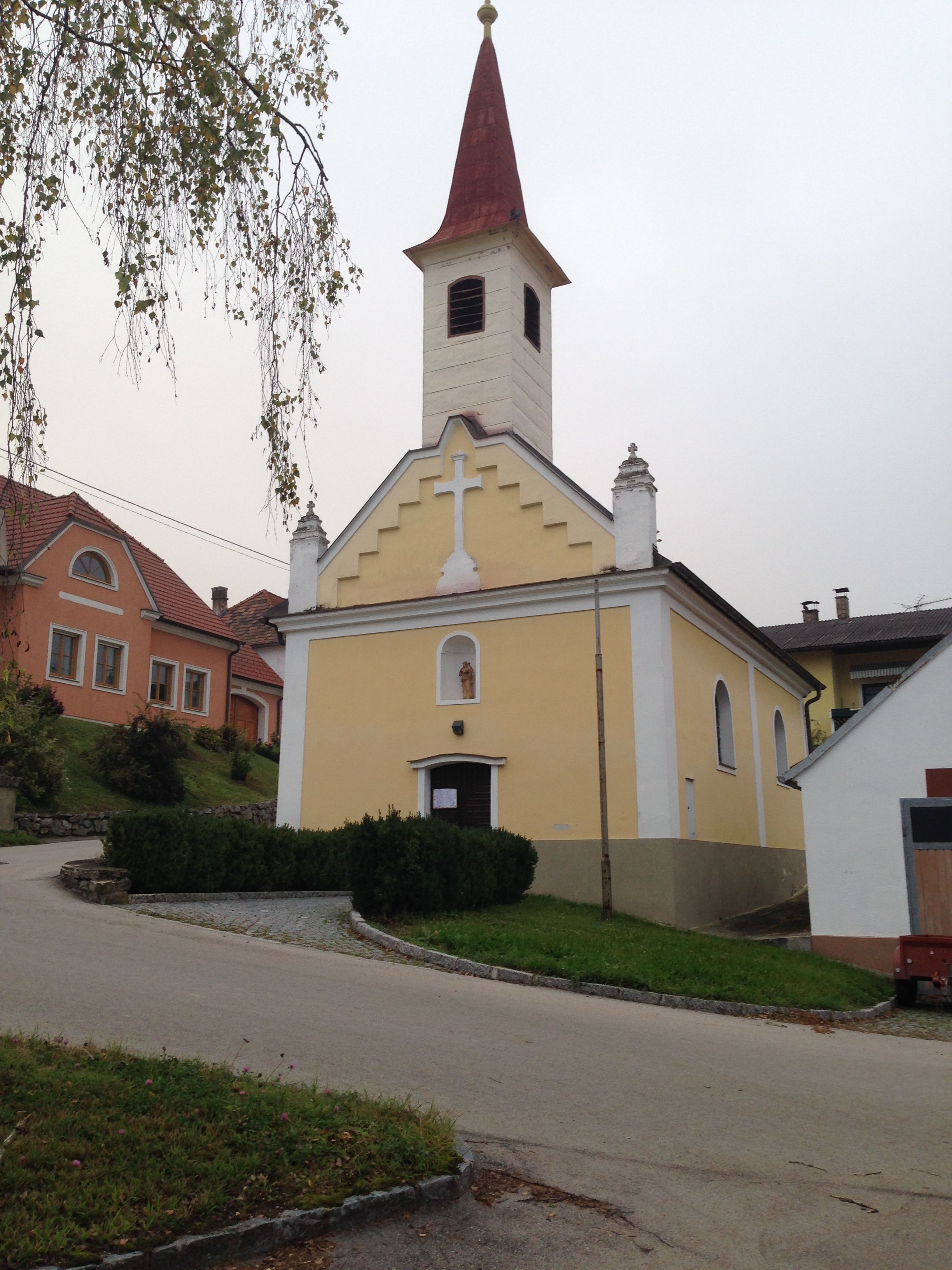 Eine kleine gelbe Kapelle mit rotem Turmdach in Engelmannsbrunn, umgeben von grünen Büschen und einem gepflasterten Weg.