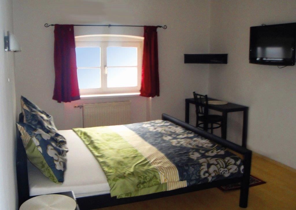 Ein Schlafzimmer mit einem Bett, einem Fenster mit roten Vorhängen, einem kleinen Tisch mit Stuhl und einem an der Wand montierten Fernseher.