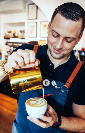 In einem einladenden Café wird mit viel Leidenschaft ein kunstvoller Cappuccino zubereitet. Der Barista lächelt, während er die cremige Milch mit geschickten Bewegungen in die Tasse gießt und ein wunderschönes Muster kreiert. Die Atmosphäre ist warm und einladend, perfekt für eine entspannte Kaffeepause.